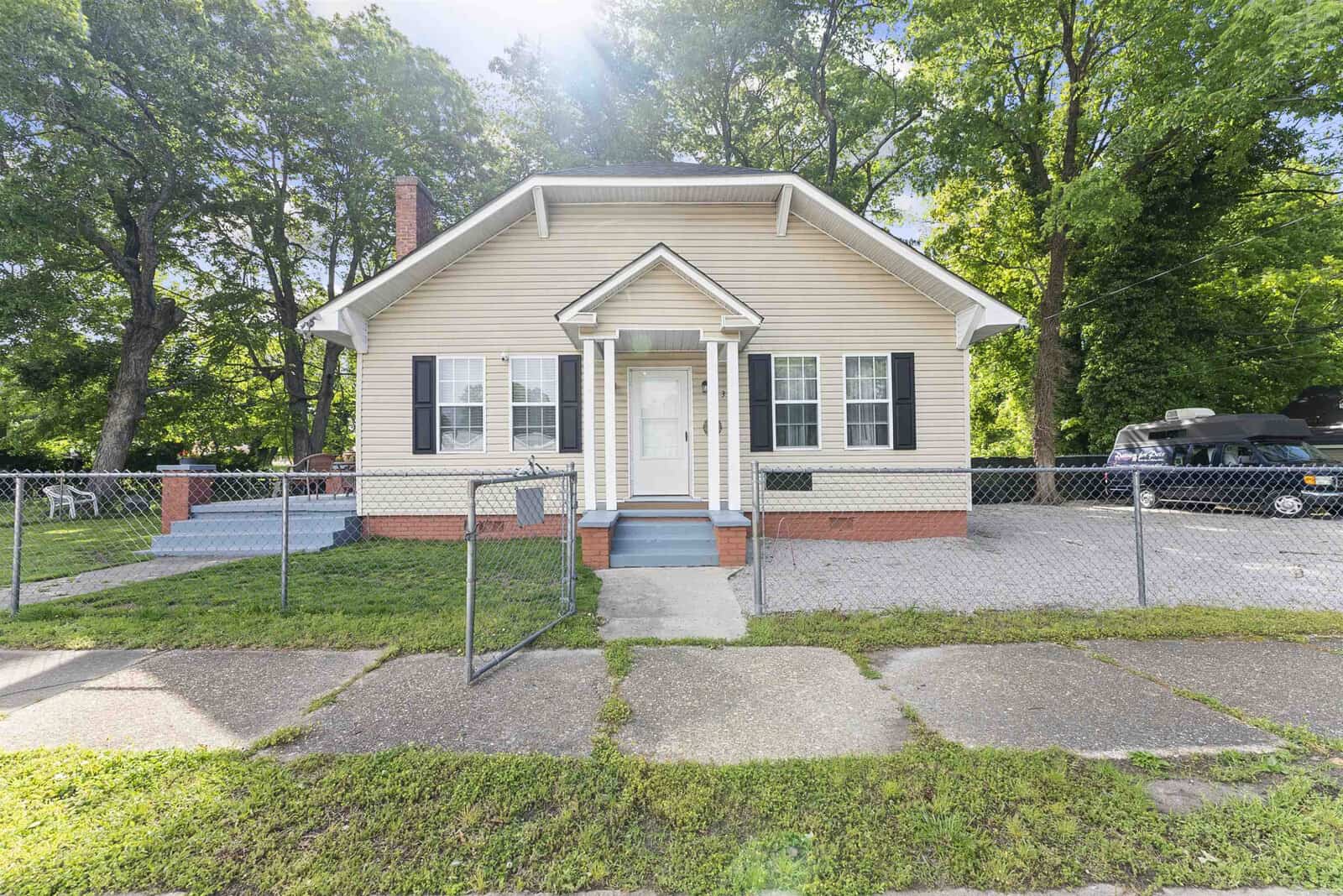 A single-story house with beige siding and white trim, featuring a centered front door and symmetrically placed windows, enclosed by a chain-link fence, with a green lawn and trees in the background.