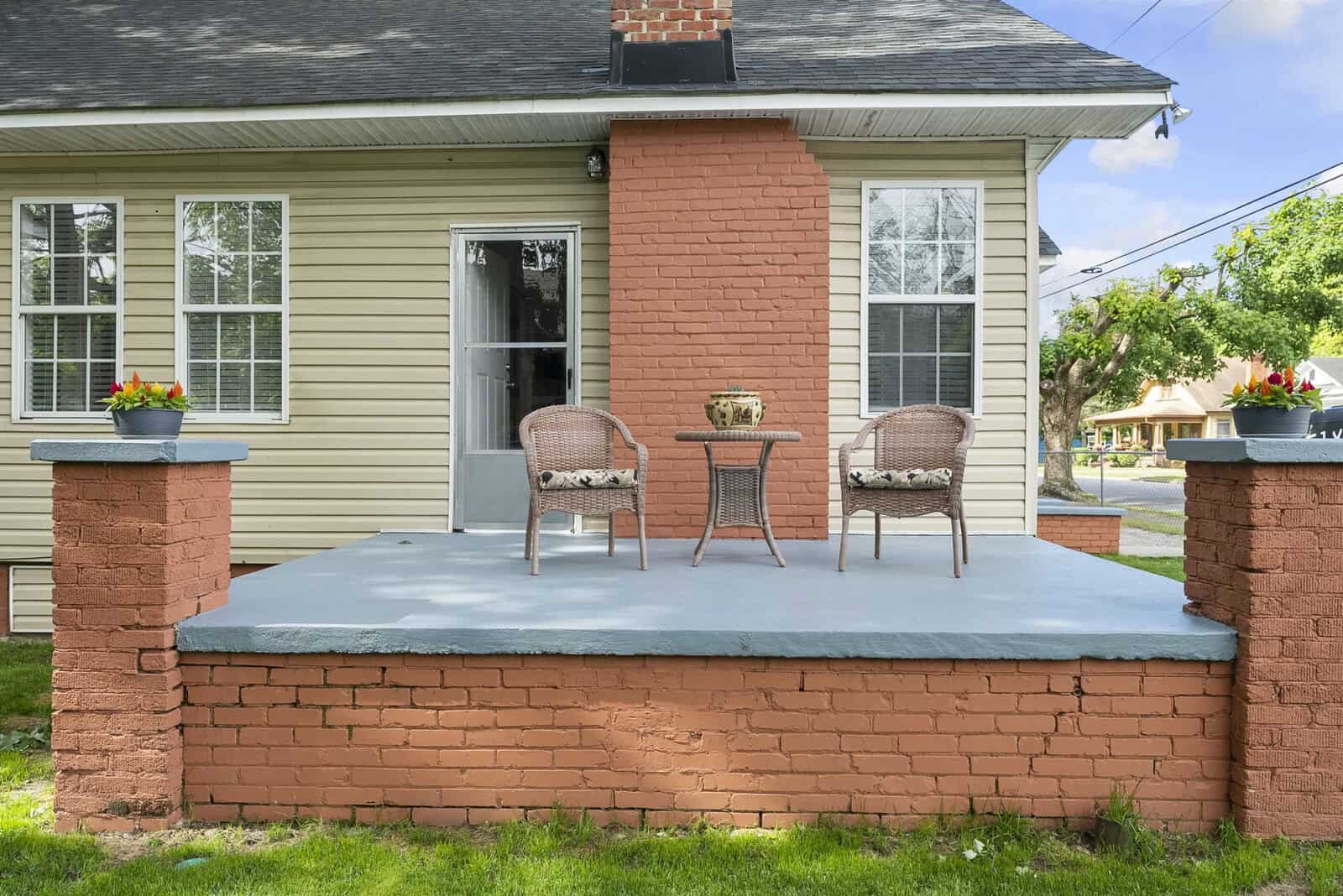 Two chairs and a table set on a concrete porch with brick pedestals, in front of a house with beige siding and a red brick chimney.