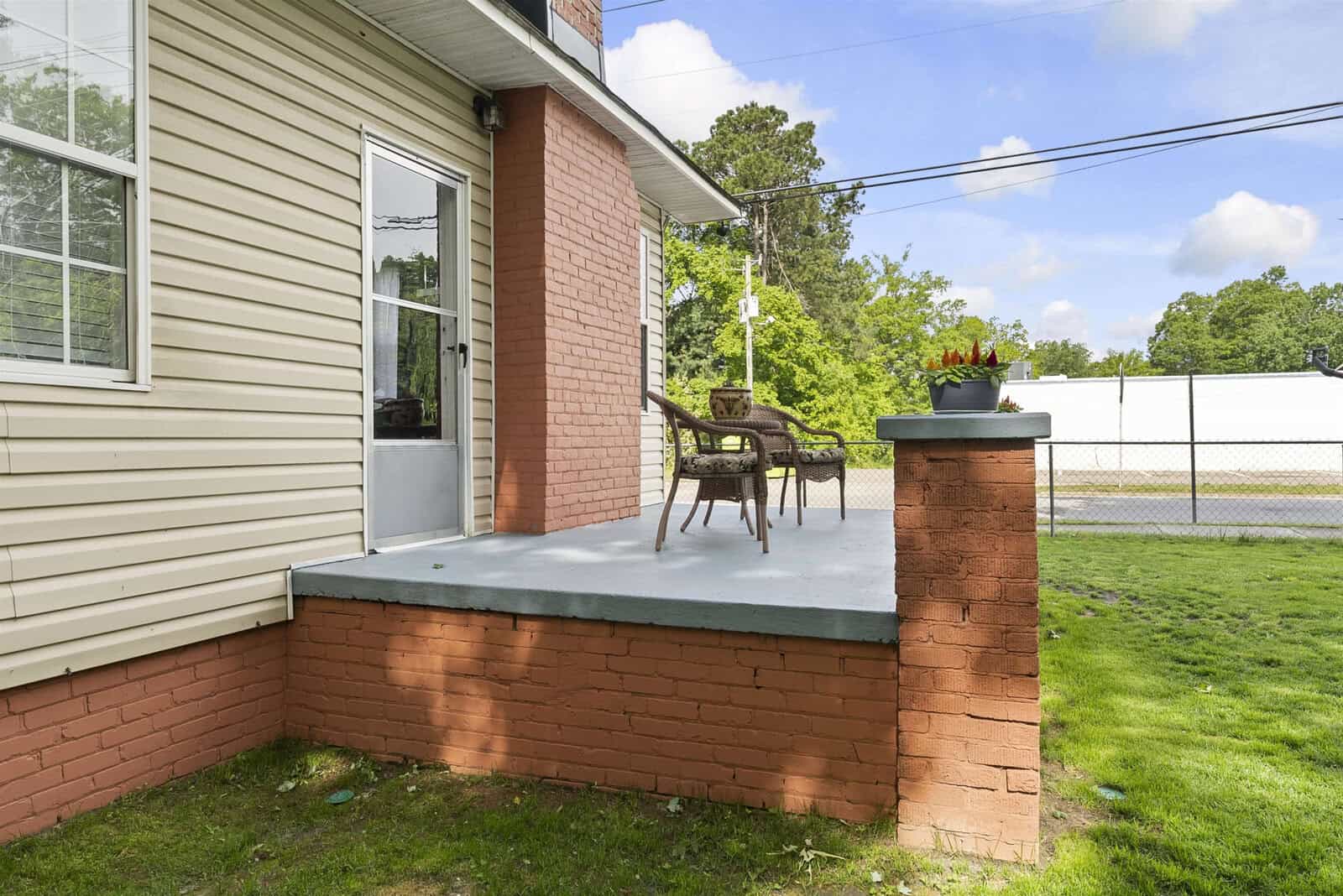 A small patio with a single chair and a table adorned with potted plants outside a brick and siding-clad house, overlooking a fenced backyard with lush green grass.