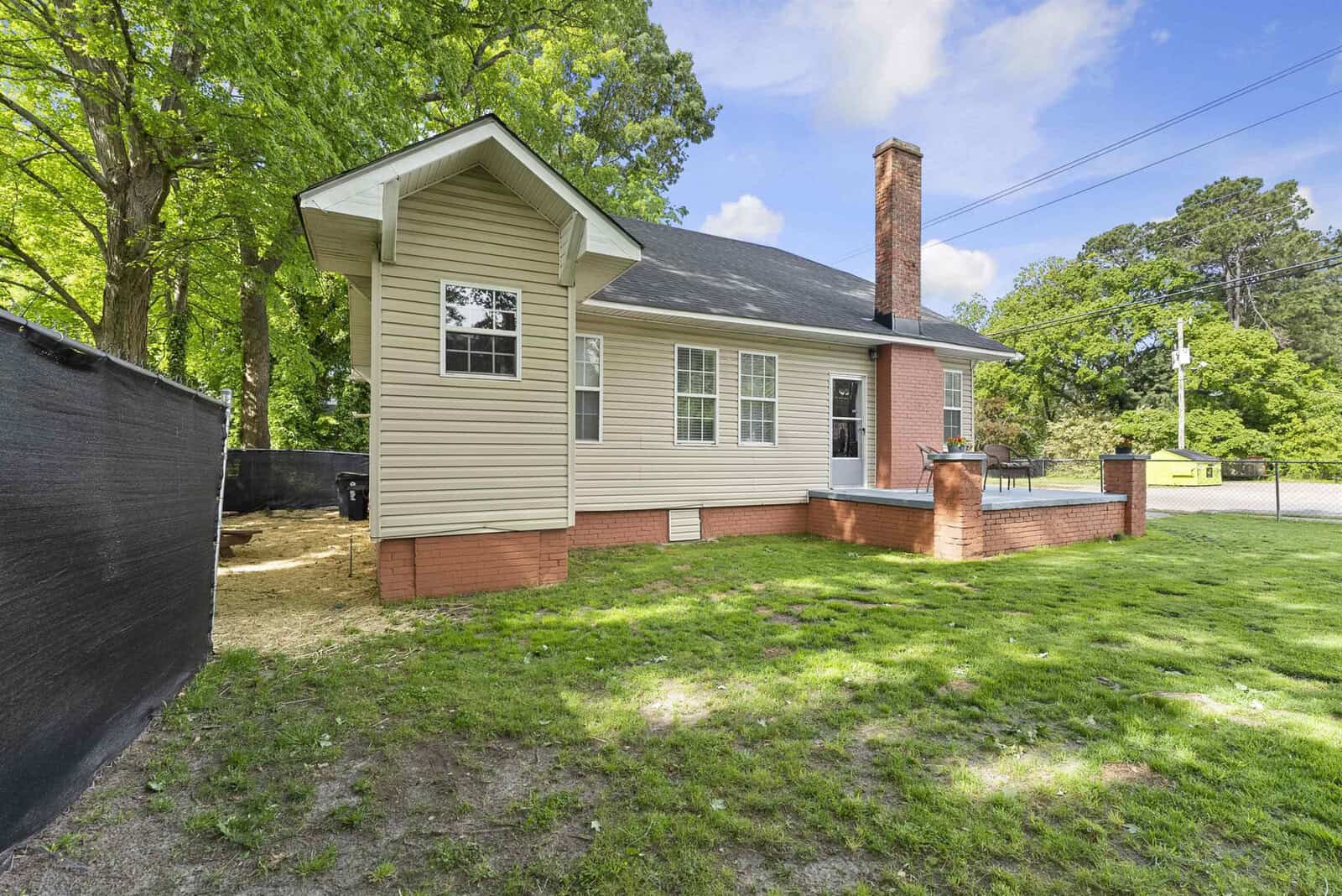 A single-story beige house with white trim, a brick chimney, and a fenced yard surrounded by trees.