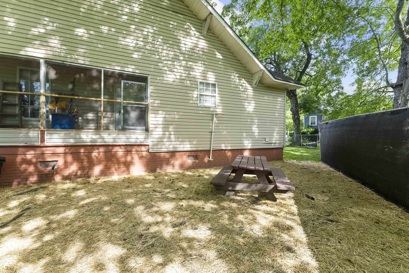 A residential backyard with freshly spread straw mulch, featuring a wooden picnic table and an above-ground pool enclosed by a black barrier, located adjacent to the side of a beige house with greenery in the background.