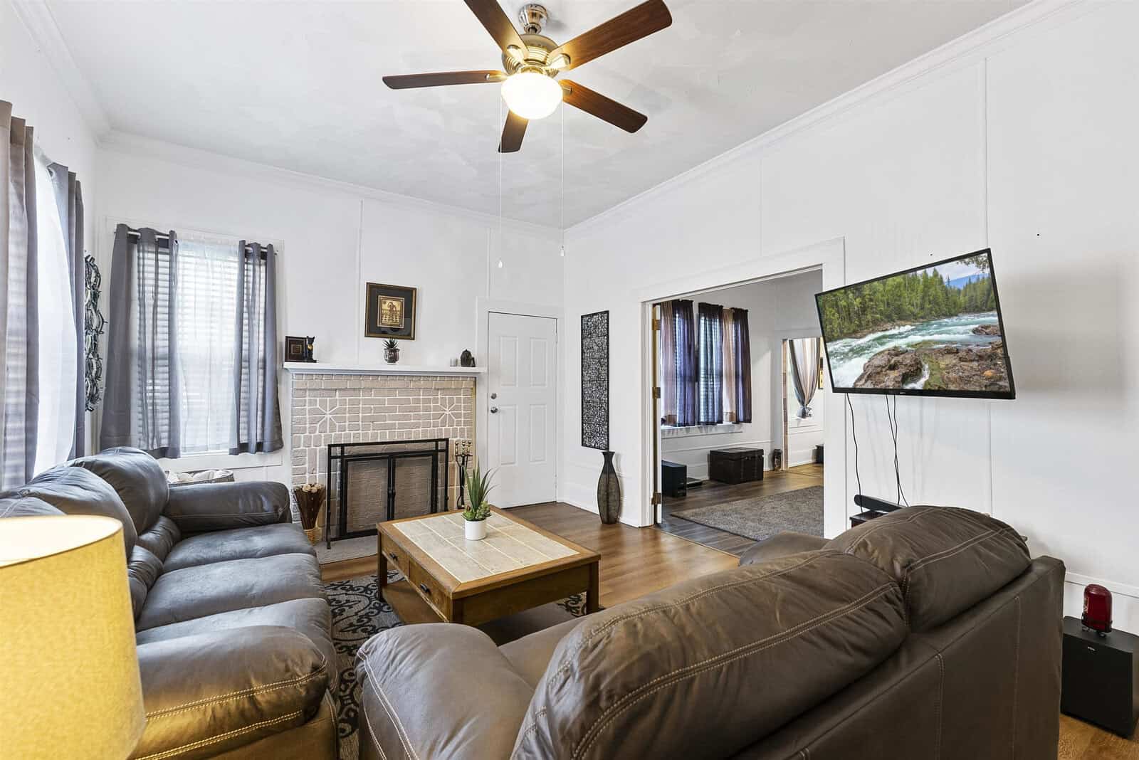 A well-lit, spacious living room featuring a cozy brown leather sofa set, a wooden coffee table, a white fireplace, and a mounted flat-screen television displaying a nature scene. the room is completed with a ceiling fan, contrasting curtains, and hardwood floors.