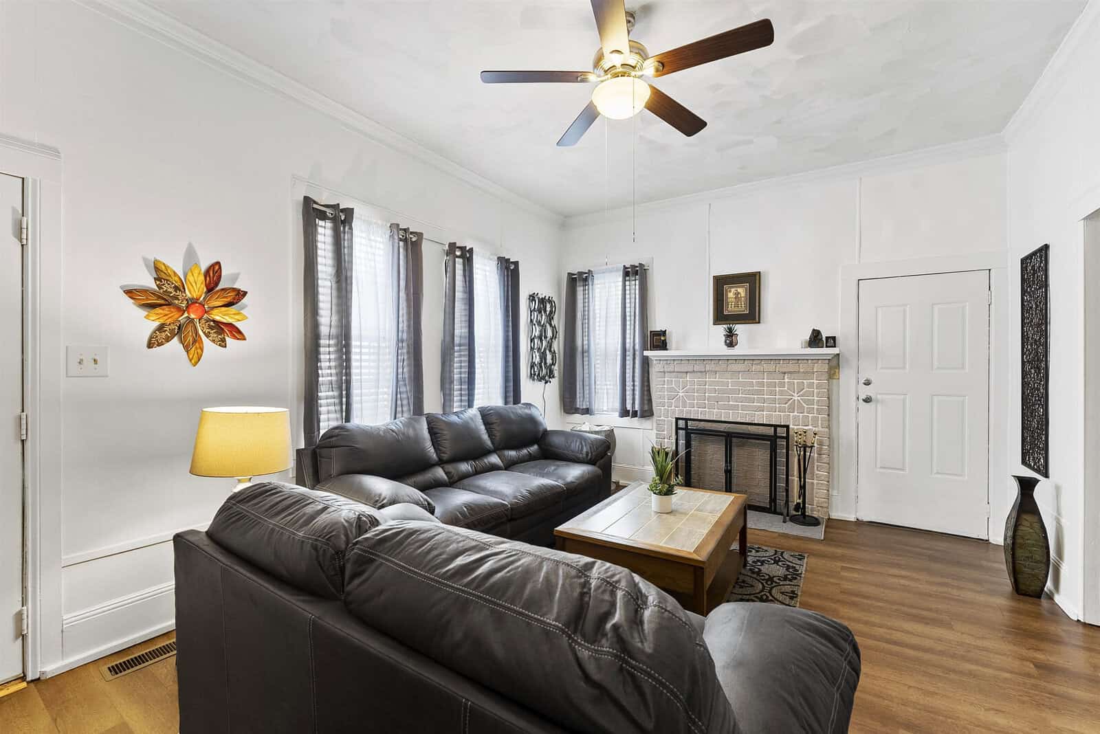 A well-lit, modern living room featuring a black leather sofa set, a wooden coffee table, a decorative fireplace, and a ceiling fan, with a colorful floral wall art adding a touch of vibrancy.