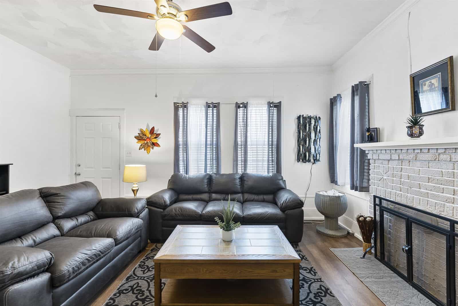 A well-lit living room with a dark leather sofa set, a central coffee table with small plants, a white fireplace, various decorative items, and a ceiling fan.