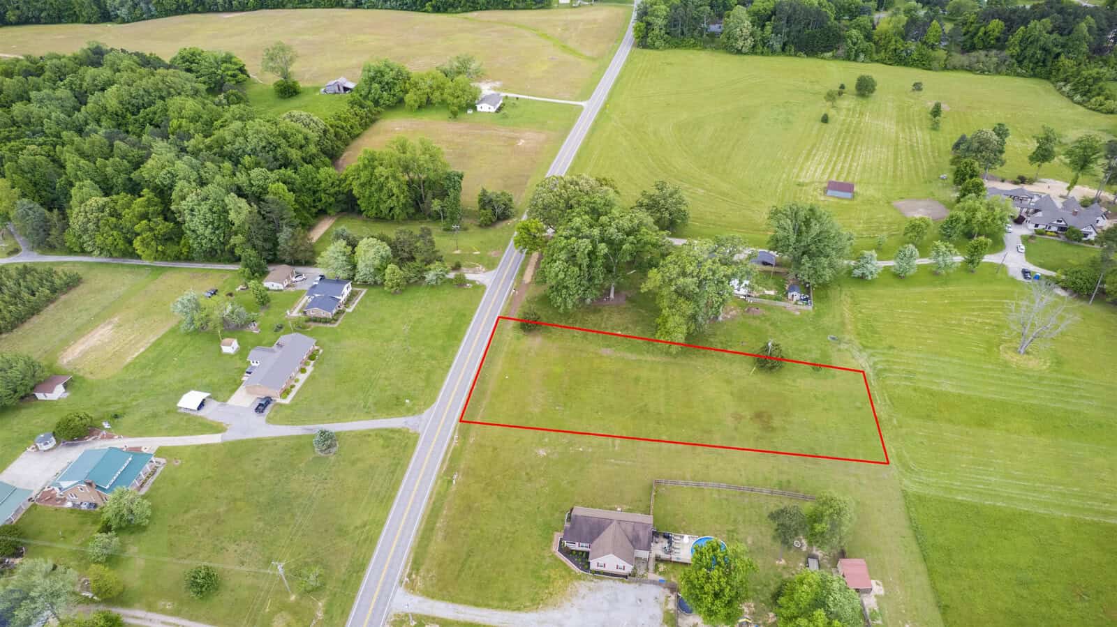 Aerial view of a rural landscape with a highlighted property boundary marked in red on a grassy field, adjacent to a road and surrounded by scattered houses and trees.