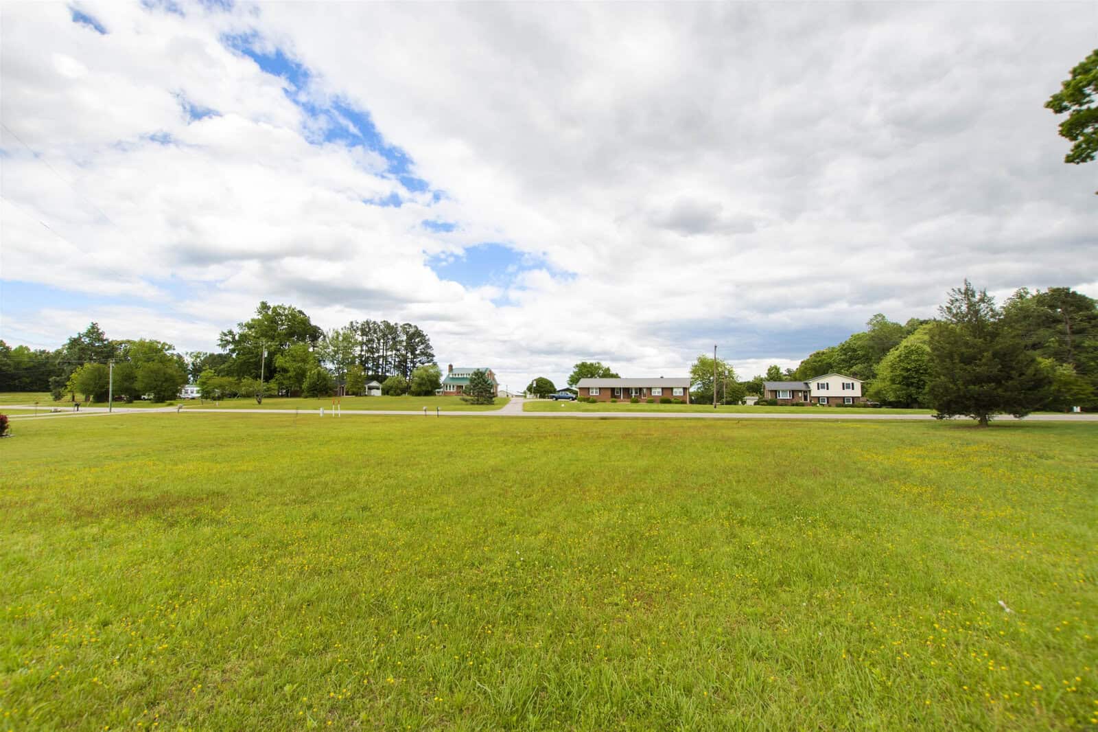 A wide-open grassy field under a partly cloudy sky, with a few buildings visible in the distance.