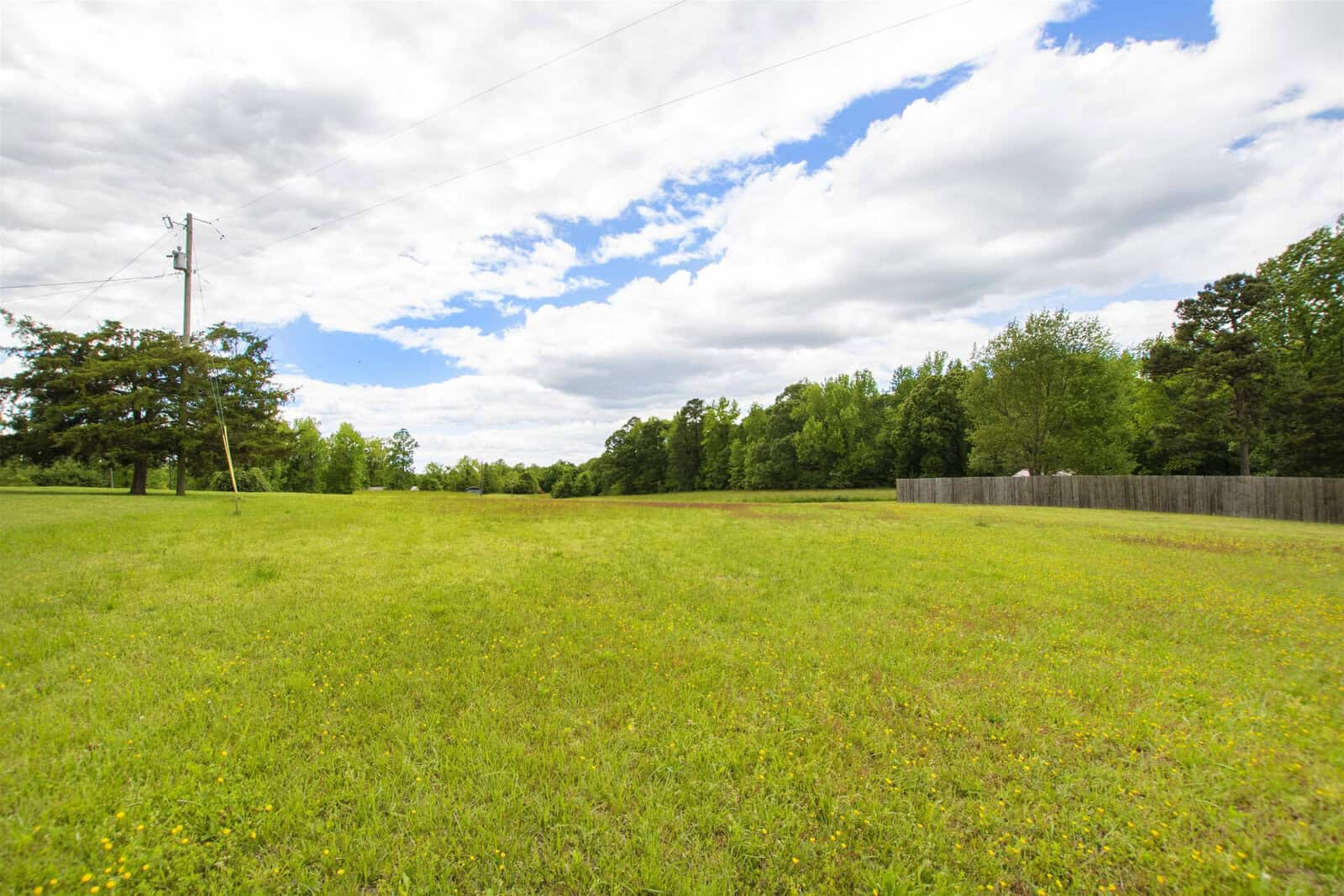 A spacious grassy field with power lines and a wooden fence bordering a forest under a partly cloudy sky.