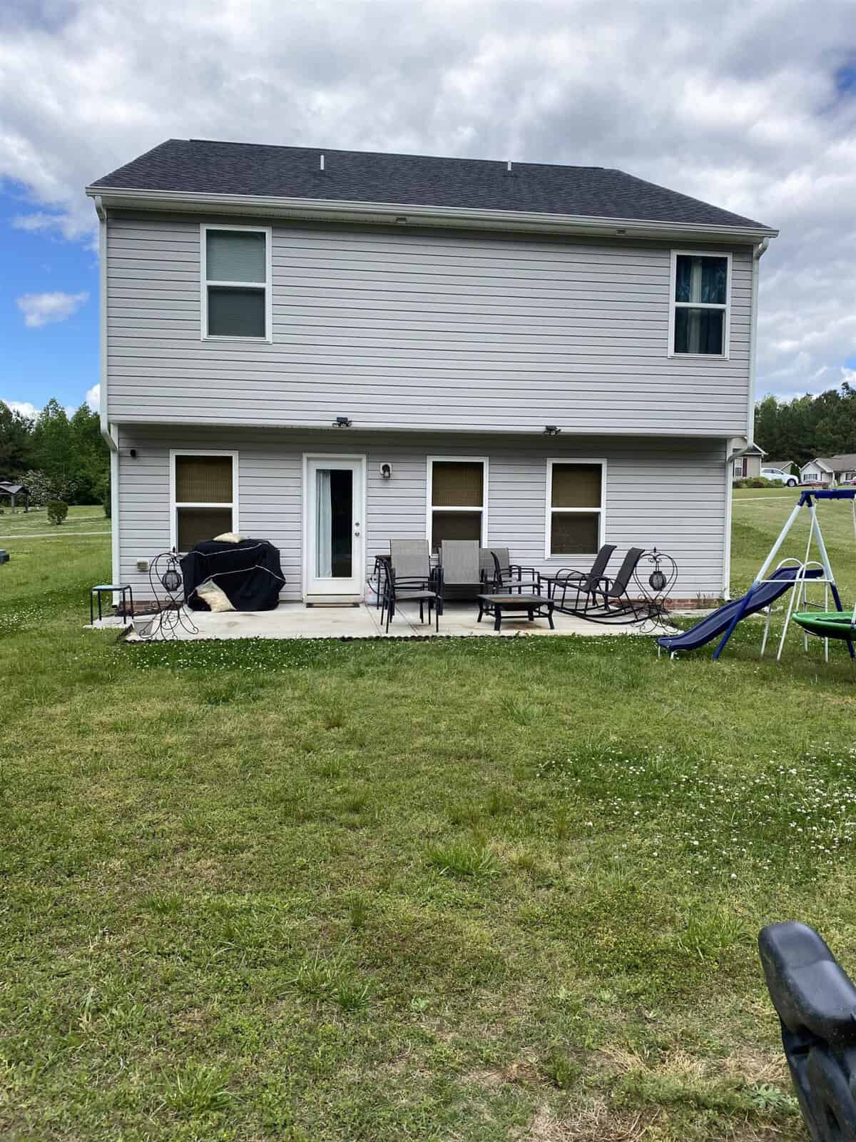 A two-story house with white siding and black shutters, featuring a backyard with a grill, outdoor furniture, and a children's slide.
