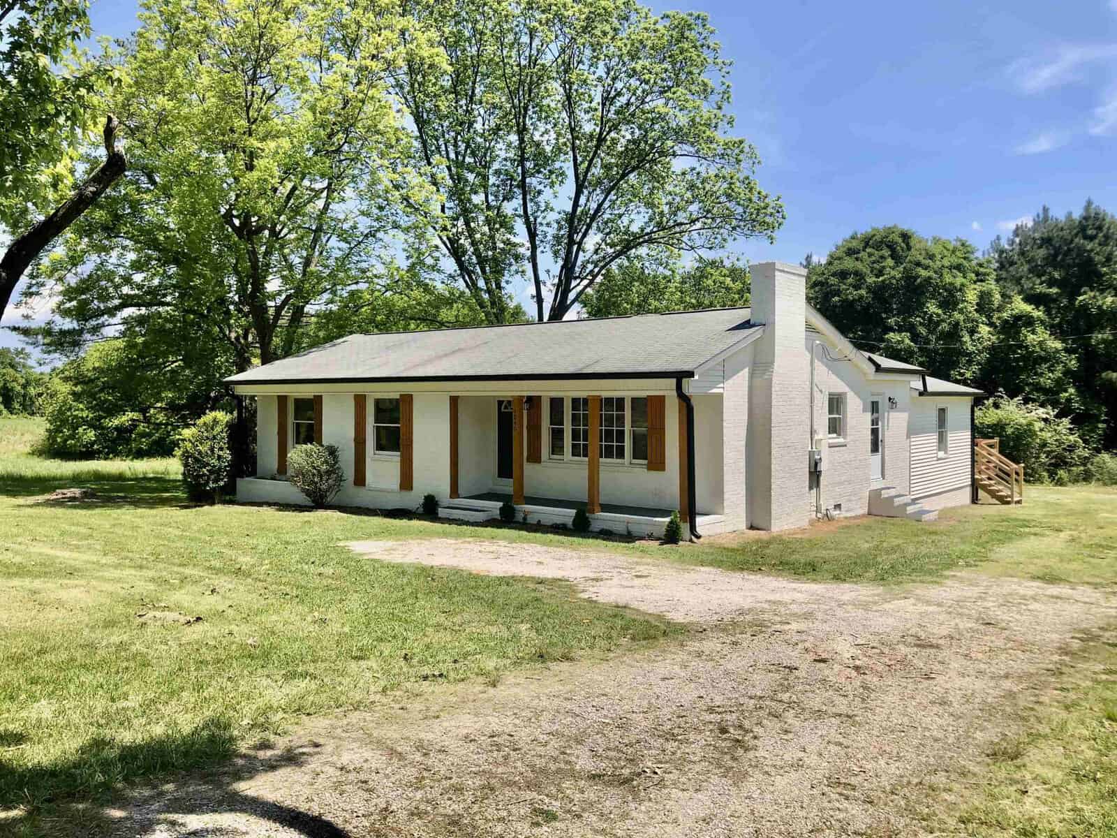 A single-story house located at 1840 Satterwhite Point Road, Henderson NC, with beige siding and white trim, featuring a small front porch and surrounded by greenery under a clear blue sky