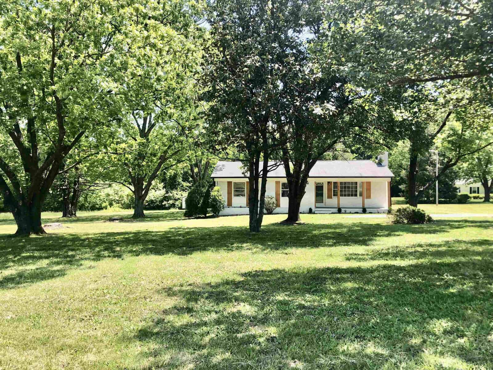 A single-story house with a yellow exterior, white trim, and a covered front porch, located in a green lawn with mature trees.