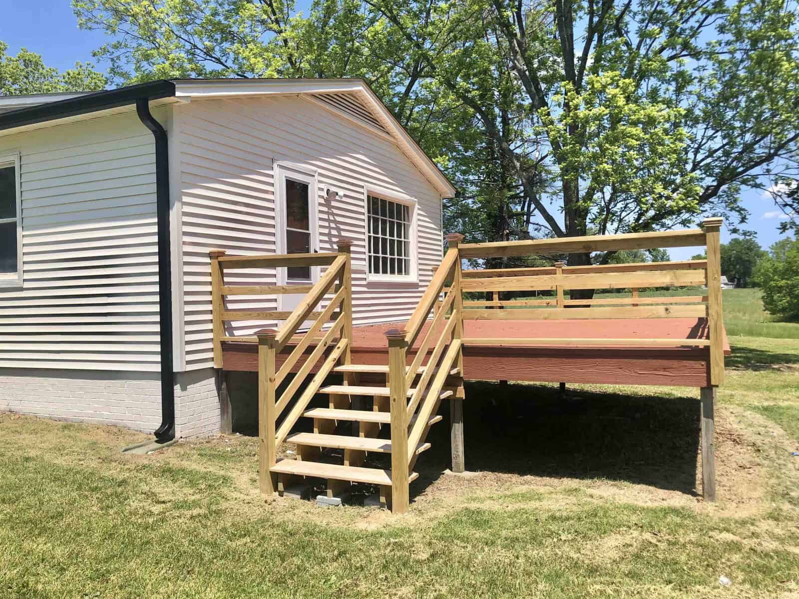 A wooden staircase leads to an elevated deck attached to the side of a light-colored mobile home, surrounded by a grassy area with trees in the background.