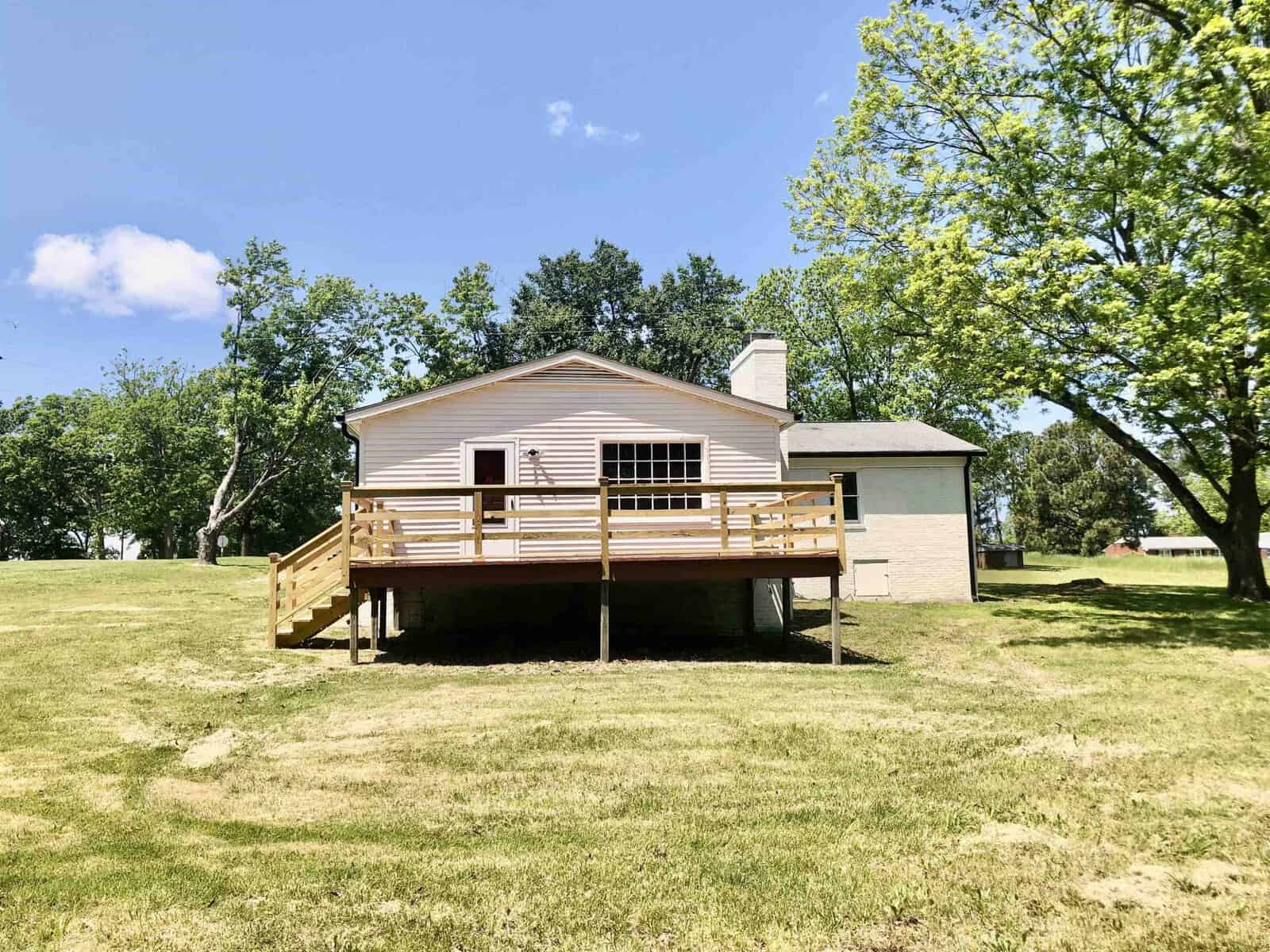 A single-story house with a wooden deck and white trim, set against a backdrop of green grass and trees under a clear blue sky.