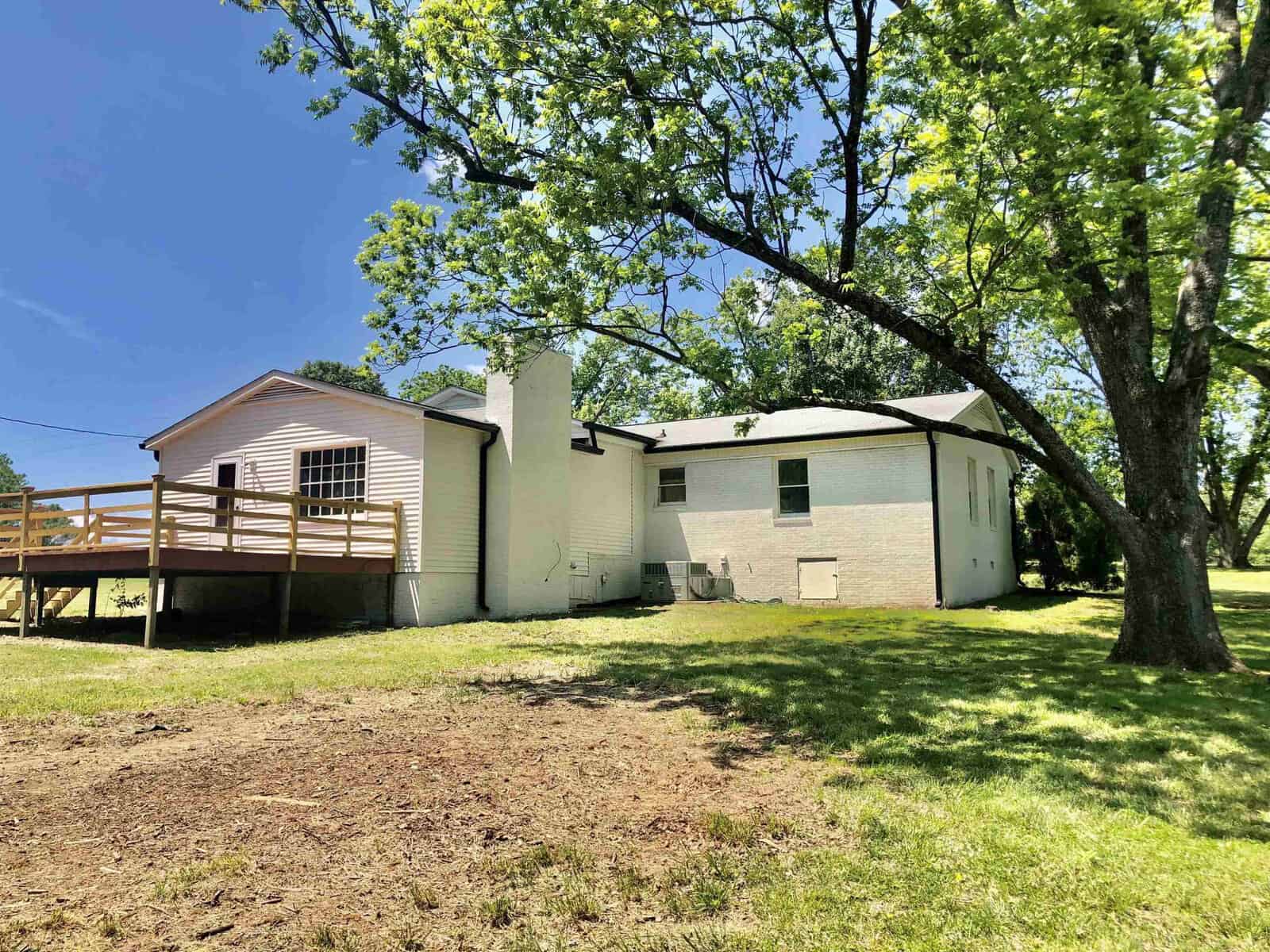 A split-level house with a beige exterior, featuring a chimney, and a wooden deck under a clear blue sky, surrounded by green trees and lawn.