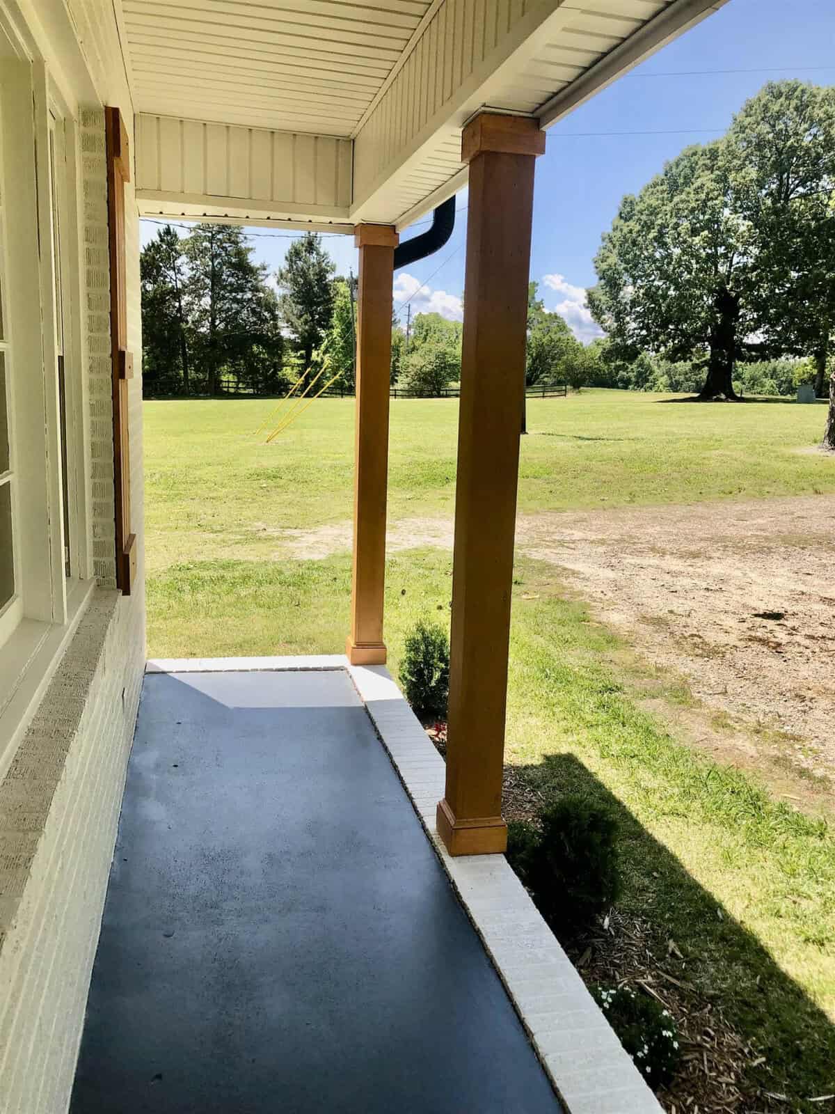 A covered residential porch with a wooden support column overlooking a grassy lawn and trees in the distance on a sunny day.