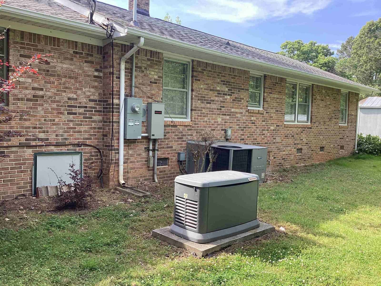 A brick house with multiple electrical boxes and a utility meter mounted on the exterior wall. In the foreground, there is a green outdoor generator unit placed on a concrete slab. The surrounding yard is grassy with some bushes near the house.