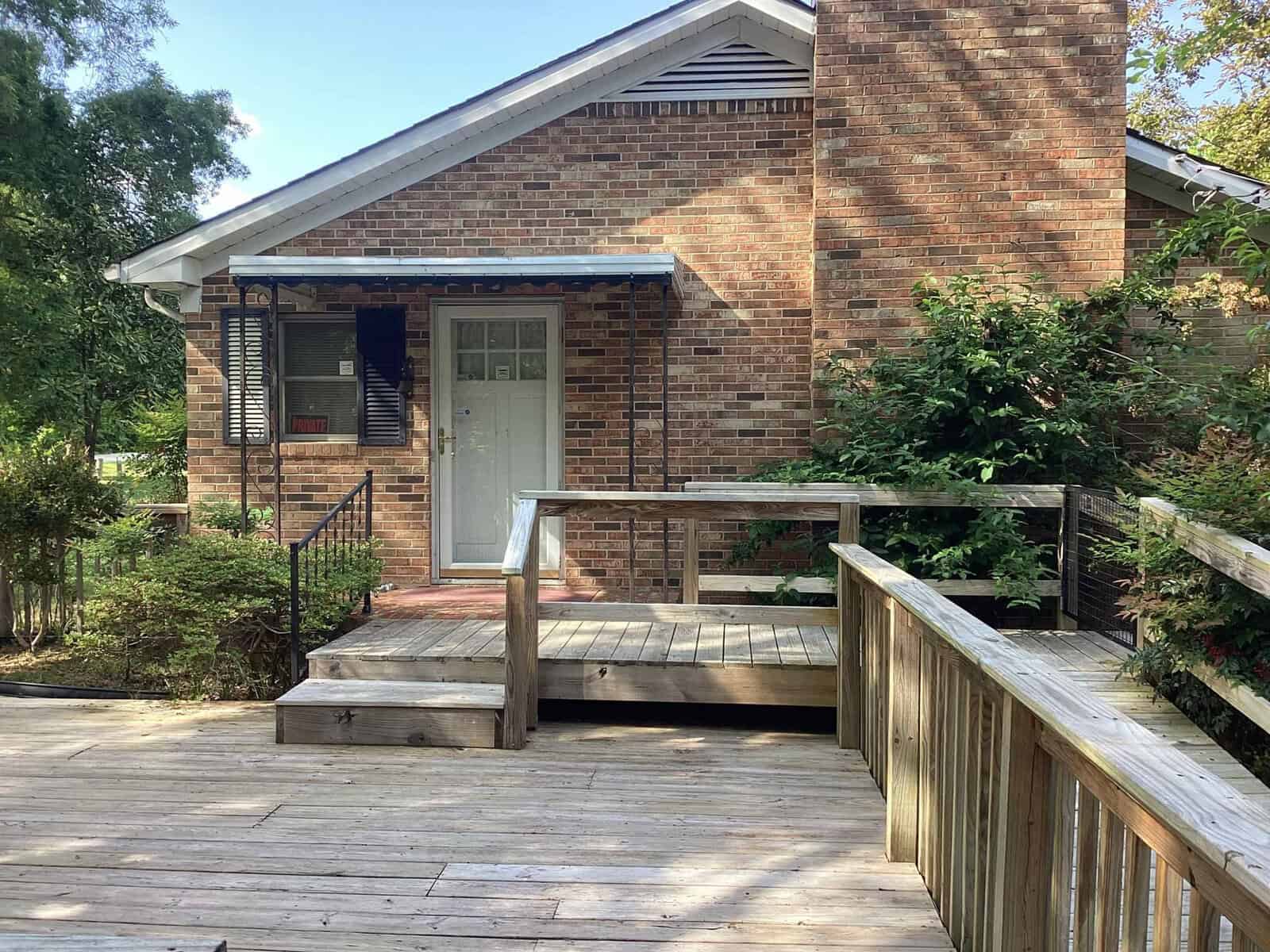 A small brick house with a white door, black shutters, and a blue awning over the entryway. A wooden ramp and steps lead up to the front door, and there is greenery on either side of the entrance. The house is surrounded by trees and bushes.