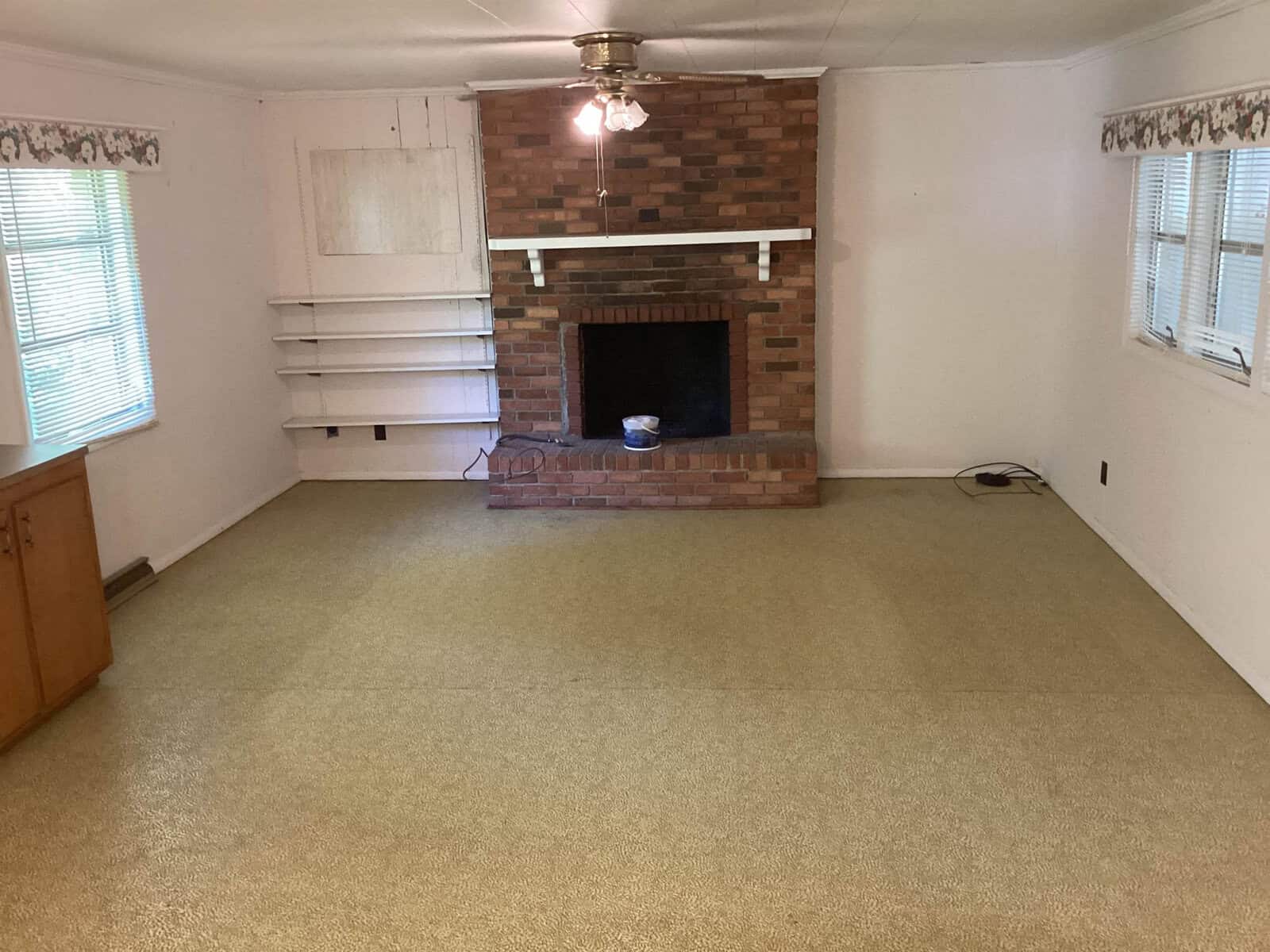 A living room with beige carpet, a brick fireplace, white shelves on the left, and a ceiling fan. There is an empty white wall above the fireplace, windows with blinds on the left and right sides, a wooden cabinet in the bottom left corner, and patterned wallpaper on top.