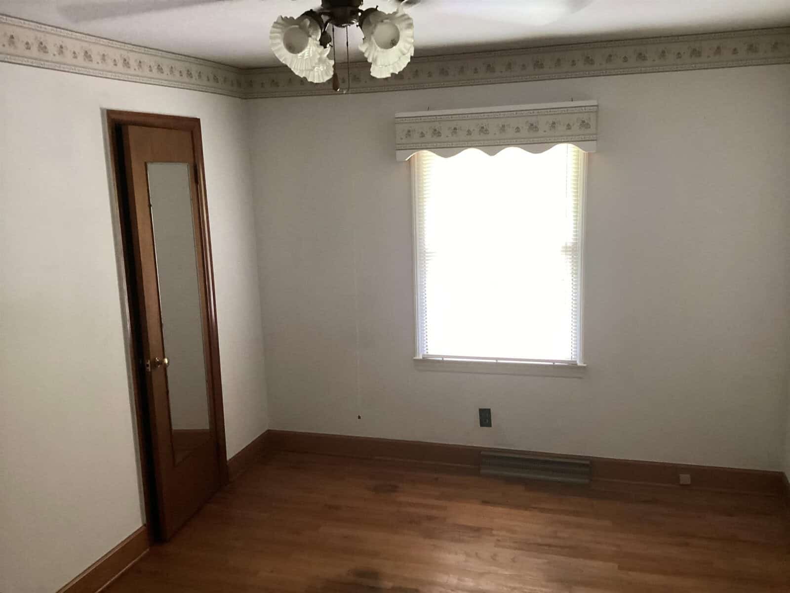 A small, empty room with hardwood floors, white walls, a wooden door with a mirror, and a window with blinds and a valance. The ceiling features a light fixture with decorative glass shades. There is baseboard trim and a vent below the window.