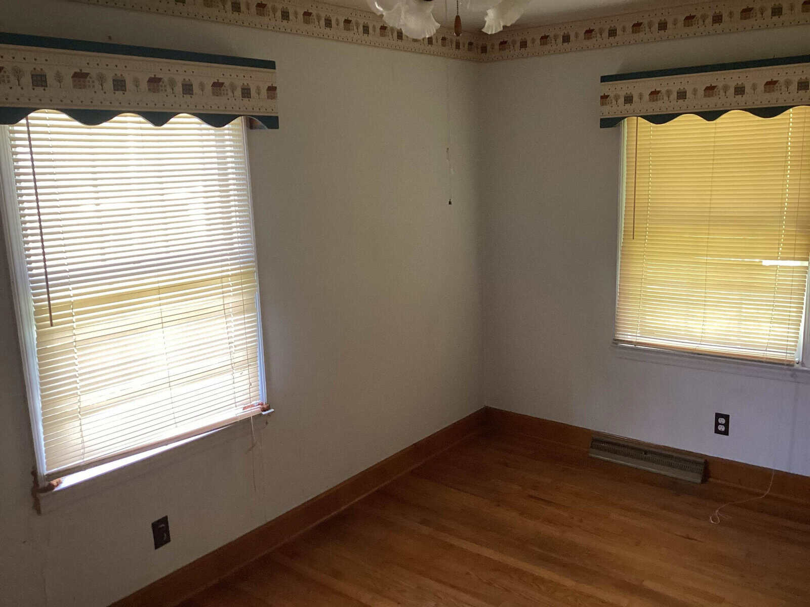 An empty room with wooden flooring and two windows with white blinds drawn halfway down. The walls are white with a decorative border near the ceiling featuring house motifs. There's a heating vent under one window and electrical outlets on the walls.