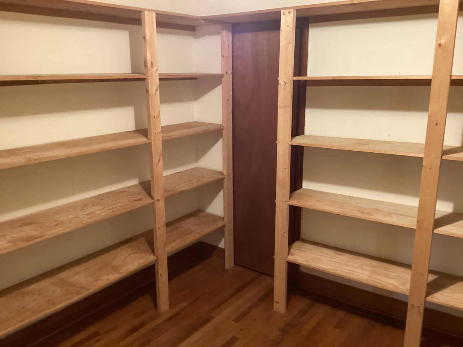 A small room featuring two sets of wooden shelves on either side, with a wooden door centered at the back wall. The shelves comprise several levels, and the room has a wooden floor. The space appears well-lit and organized, ideal for storage or a pantry.