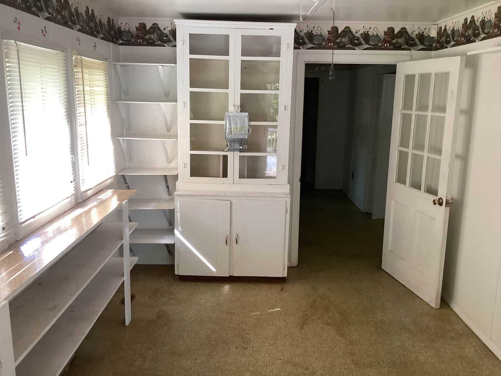 A room with light-colored walls and shelves. There are windows with blinds to the left, a white cabinet in the center with glass doors on the upper half, and an open door to the right leading to another room. The floor is carpeted.