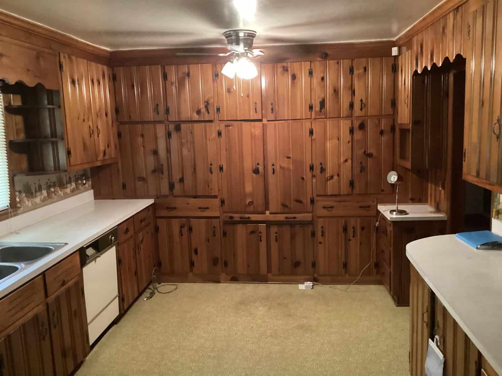 A wooden kitchen with extensive cabinetry, adorned with rustic handles and a scalloped trim along the upper edges. The room includes a sink, dishwasher, and countertop space, all under a ceiling fan with a light. The floor has a light-colored carpet.