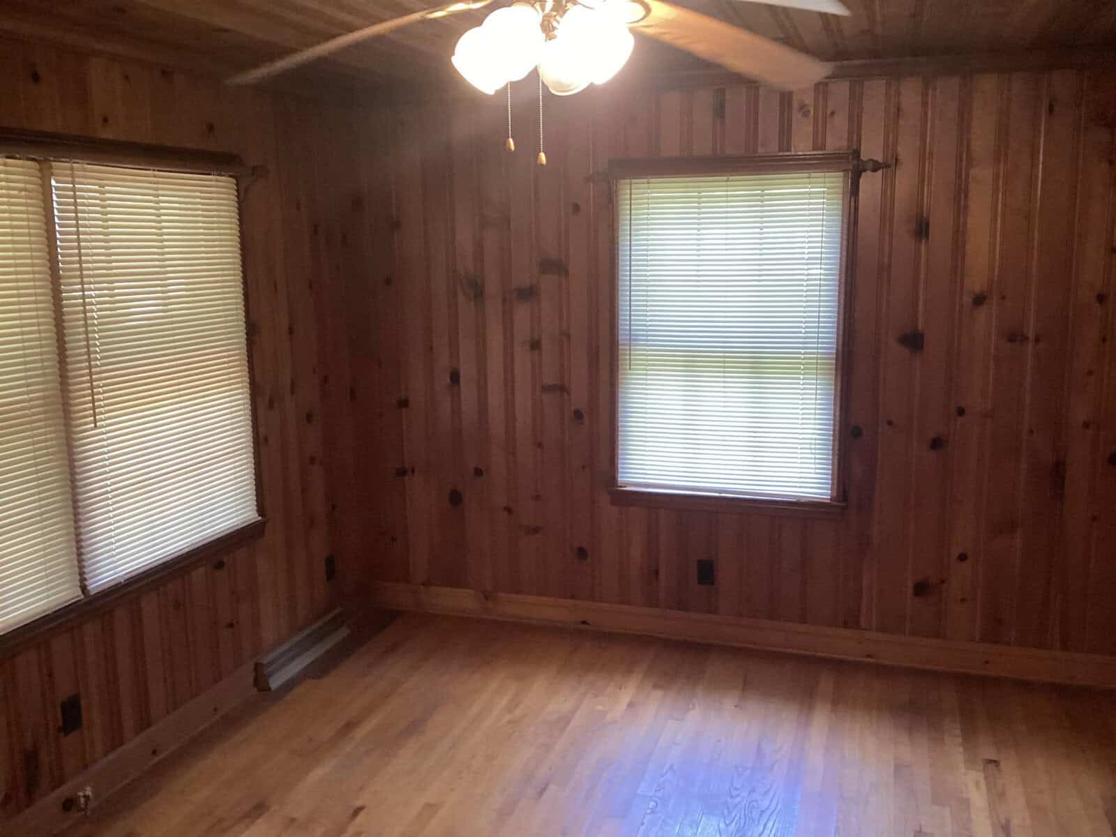 An empty room with wooden walls and hardwood floors, featuring two windows with closed blinds. A ceiling fan with lights is mounted on the wooden ceiling. The room is illuminated with natural light filtering through the blinds.