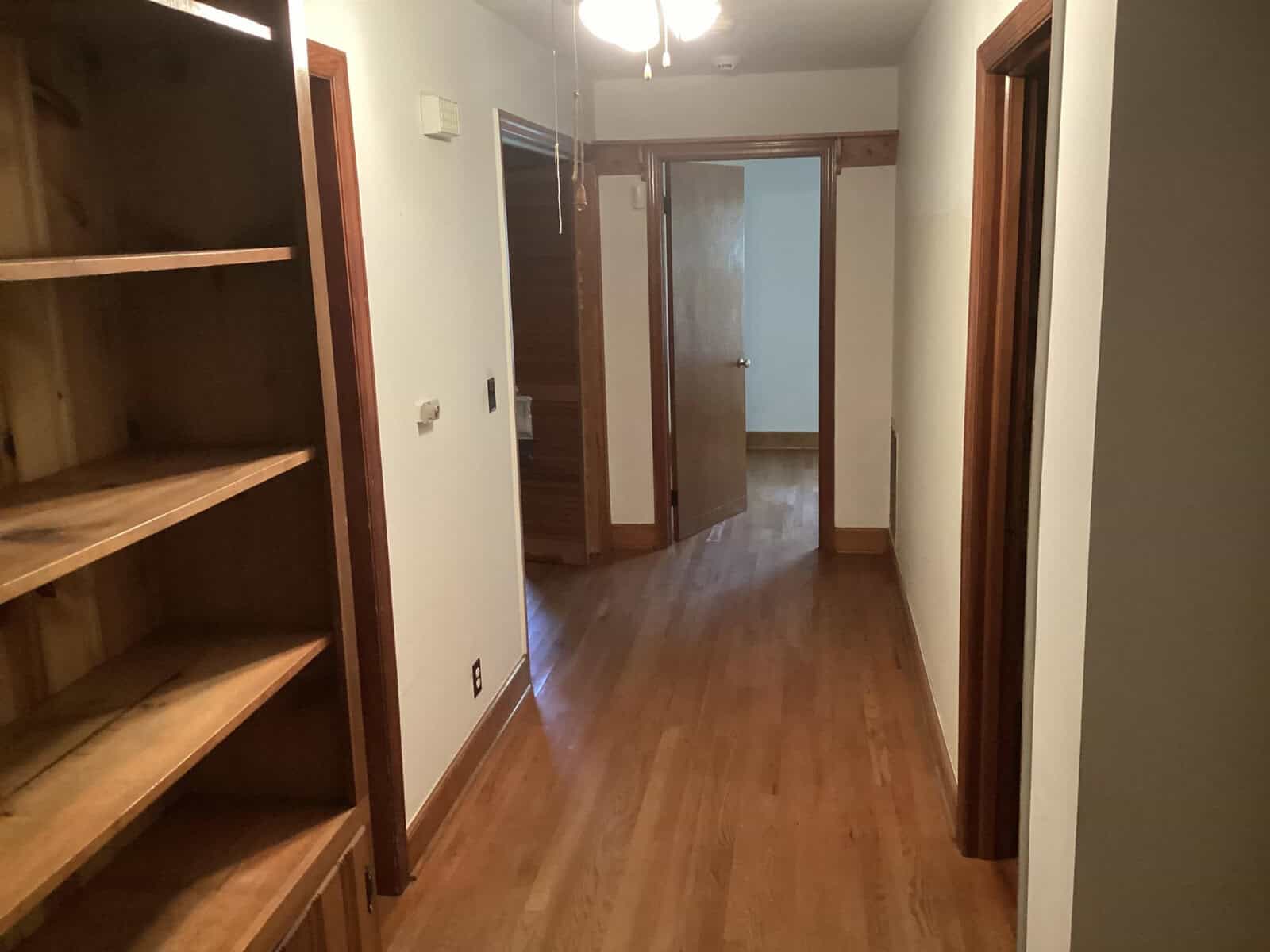 A hallway with wooden flooring and white walls. On the left side, there's a built-in wooden shelf. The hallway leads to an open door at the end, with other doors on either side. A ceiling fan is visible overhead.