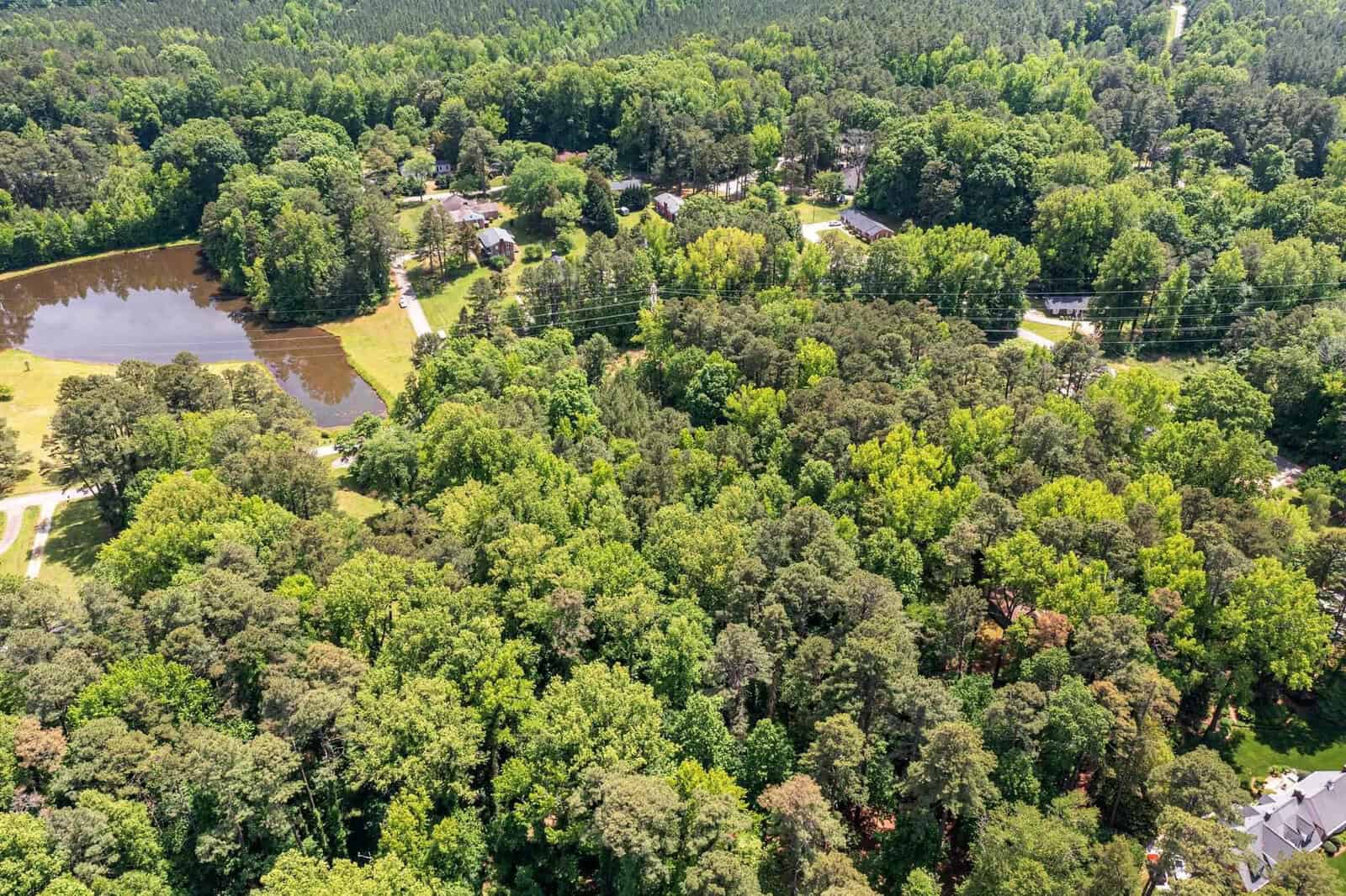 Aerial view of a residential area with houses interspersed among green trees, featuring a pond and roadways.