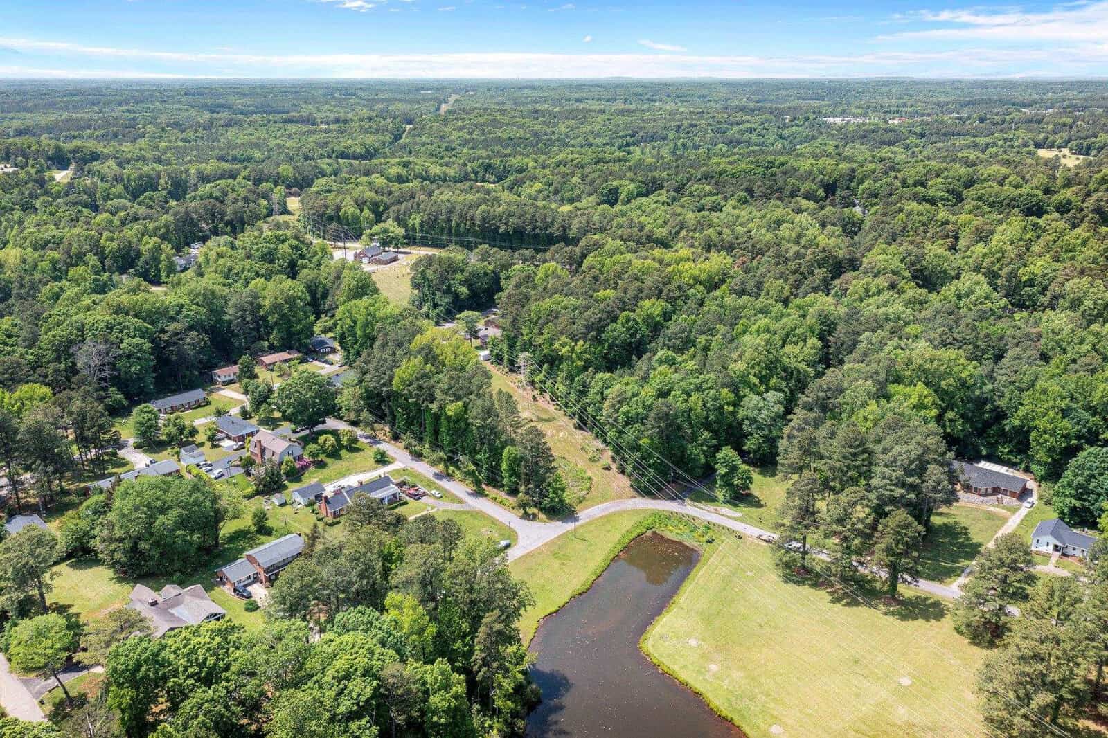 Aerial view of a residential area with houses scattered among greenery, a road winding through the center, and a body of water on the left.
