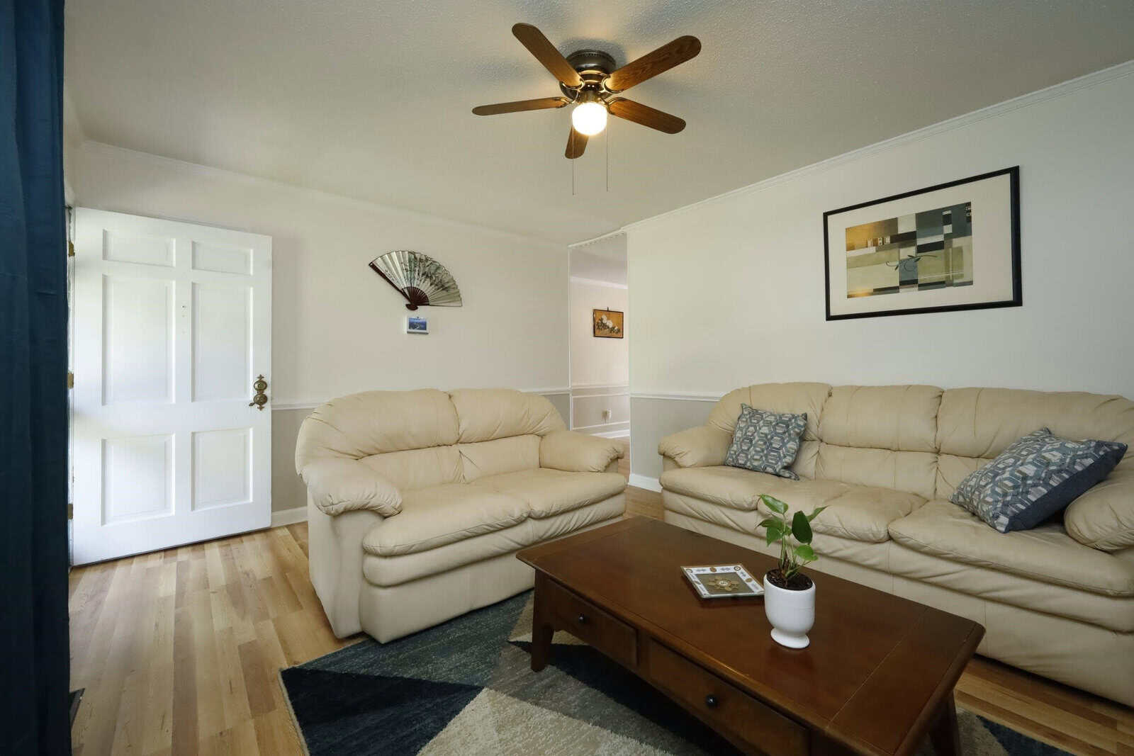 A neatly arranged living room featuring a two-piece beige sofa set with contrasting pillows, a wooden coffee table with a small plant and a magazine, a ceiling fan above, and a wall-mounted decorative fan and framed art. the room has hardwood flooring and a white door on the far end.