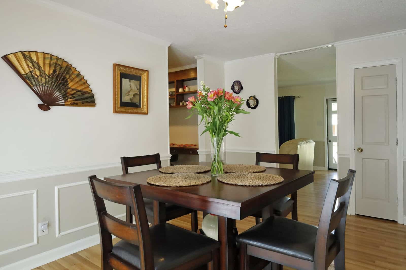 A well-lit dining room furnished with a dark wood table and chairs, decorated with wall-mounted plates, a large fan, and artwork, featuring a vase of pink flowers as a centerpiece.