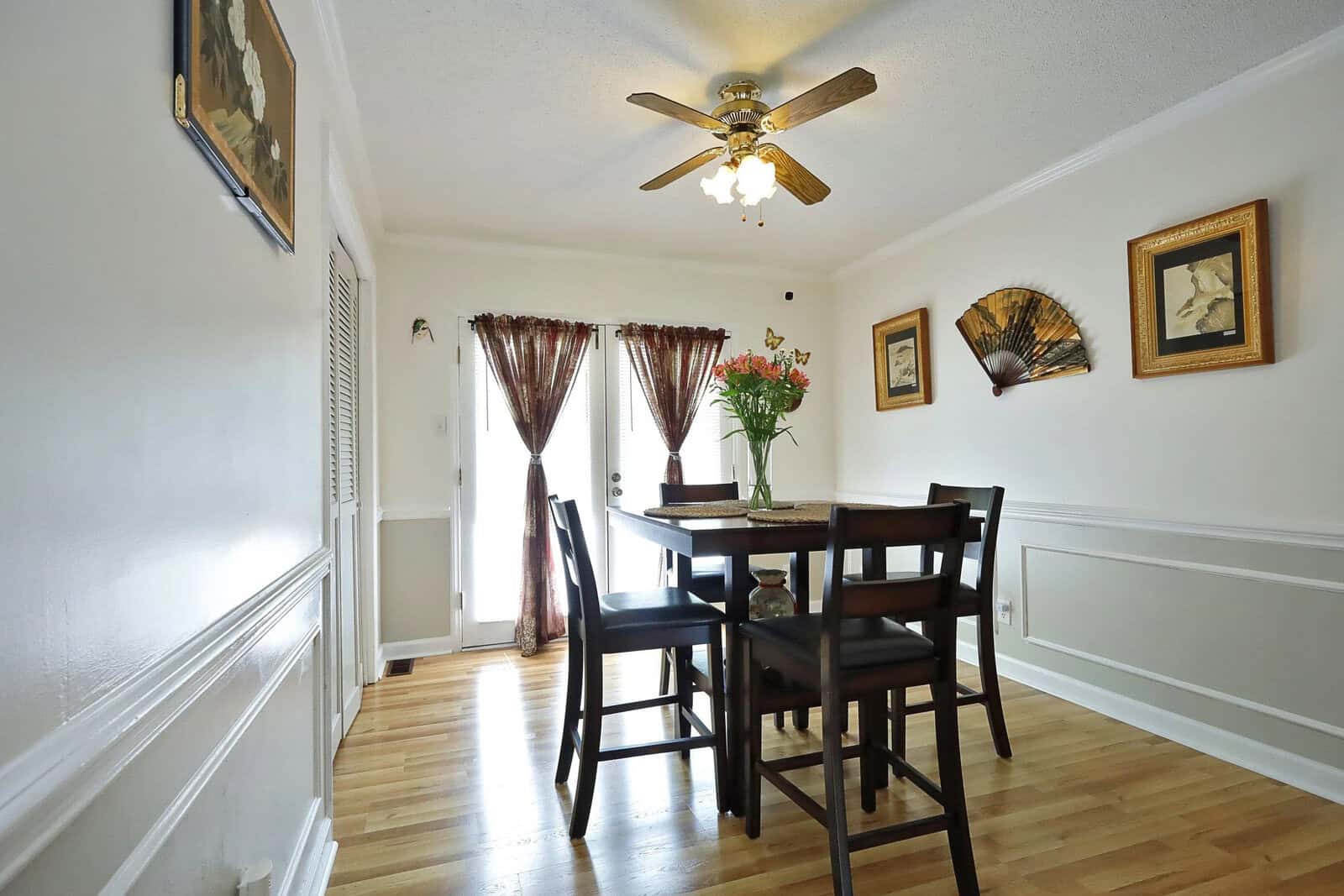 A well-lit dining room with a wooden table set for four, a ceiling fan above, and decorative elements such as wall paintings and curtains.
