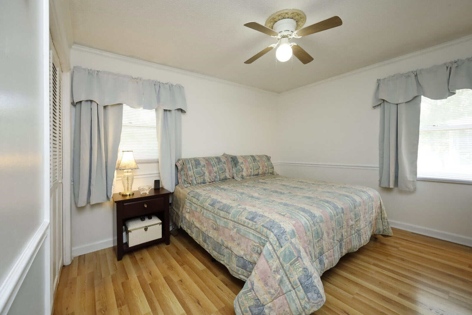 A modestly furnished bedroom featuring a single bed with a patterned bedspread, two windows with light grey curtains, a ceiling fan, and a wooden floor. a bedside table with a lamp is situated to the left of the bed.