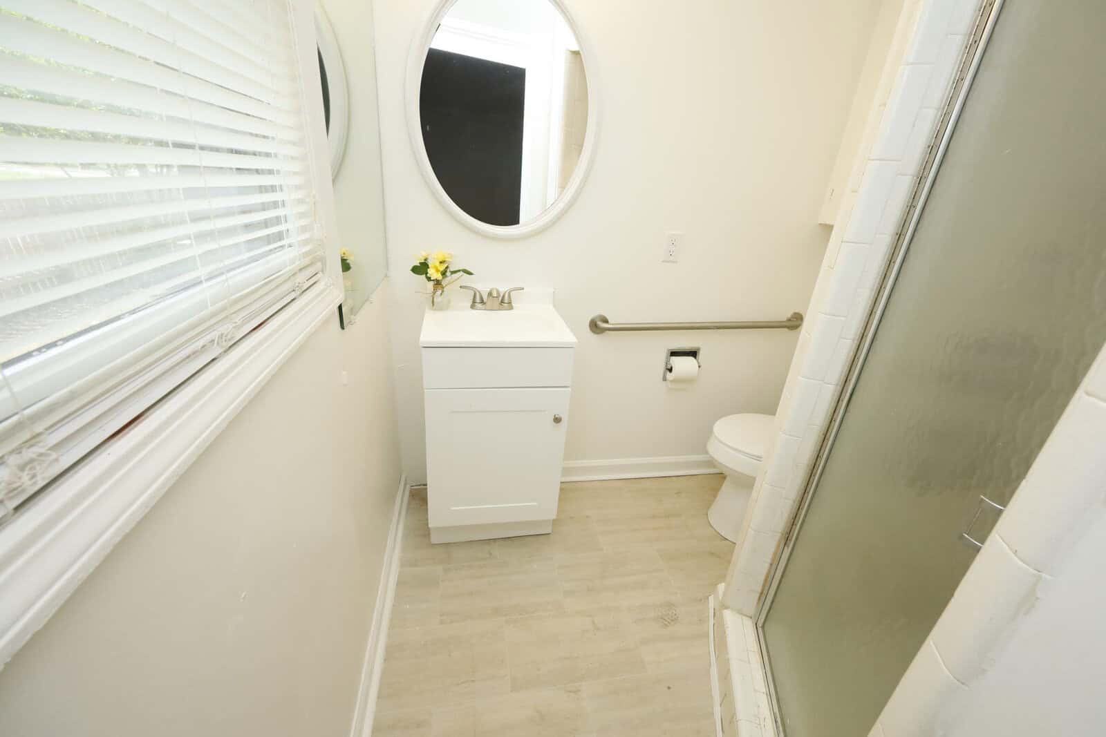 A small bathroom with a white vanity, round mirror, toilet, and a window with blinds.
