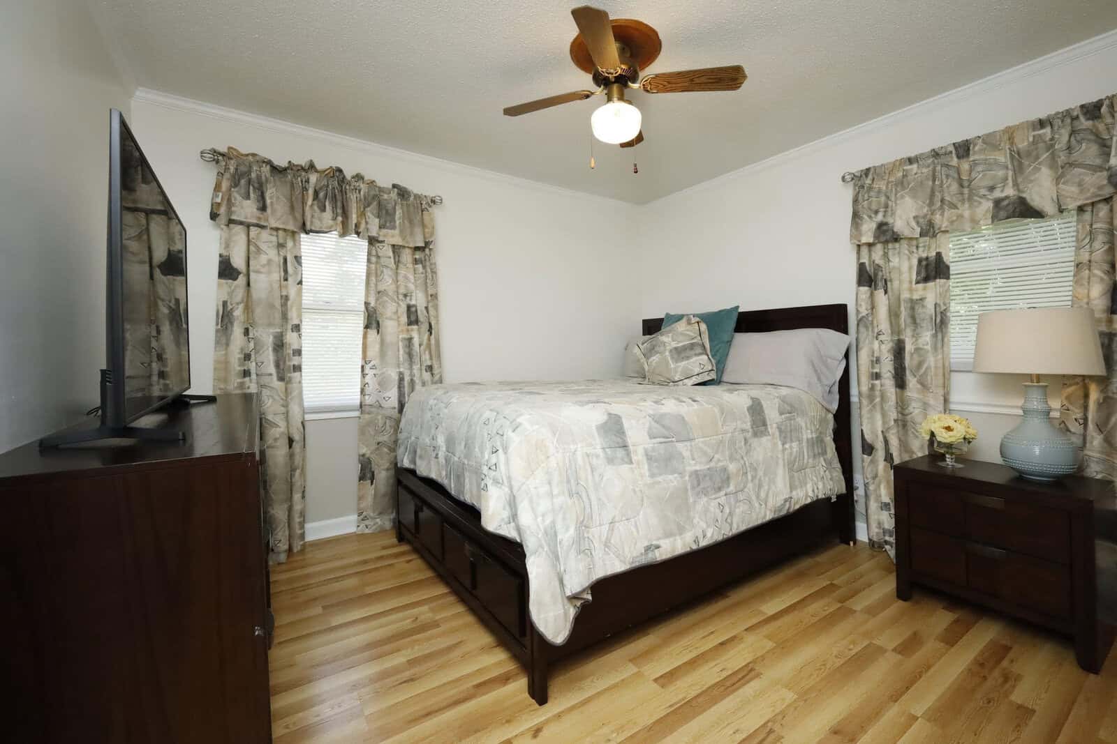 A neatly arranged bedroom featuring a made bed with patterned bedding, matching curtains, a ceiling fan, hardwood floors, a nightstand with a lamp, and a dresser with a television on top.