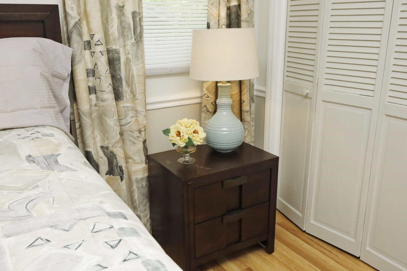 A neatly arranged bedroom with a dark wooden bedside table holding a lamp and a small vase with yellow flowers, adjacent to a bed with patterned bedding, facing window shutters.