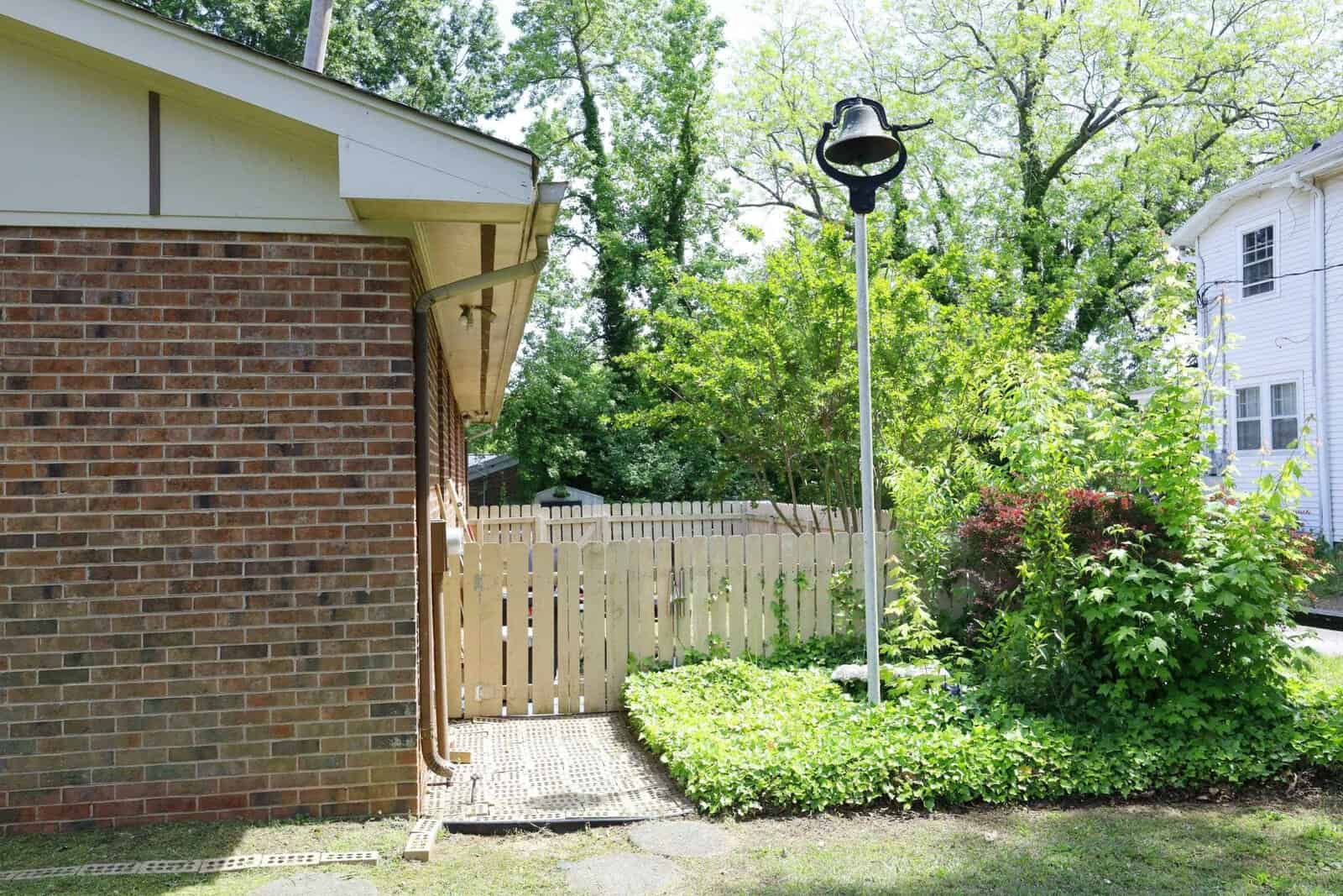 A suburban outdoor area with a brick building, wooden fence gate, lamp post, and a variety of green plants under bright daylight.