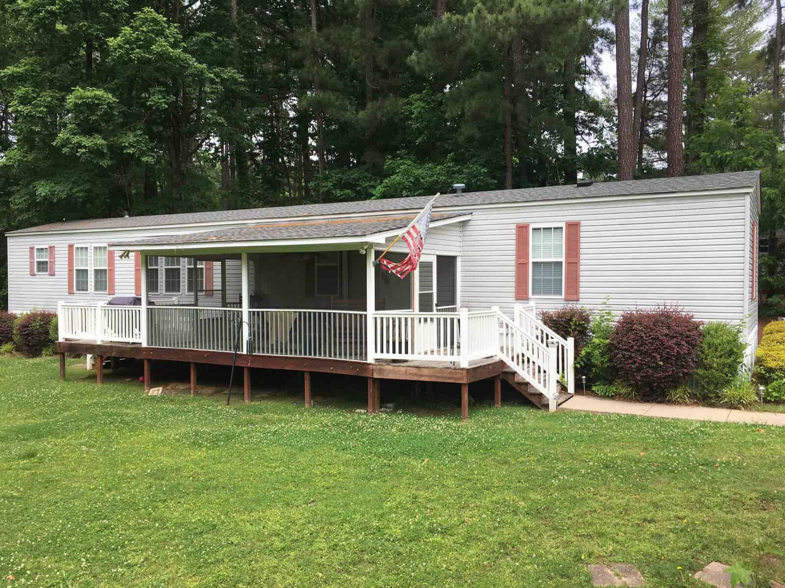 A white mobile home with a covered porch, red shutters, and an American flag is set against a backdrop of tall trees. The porch has white railings and stairs leading to a grassy yard.