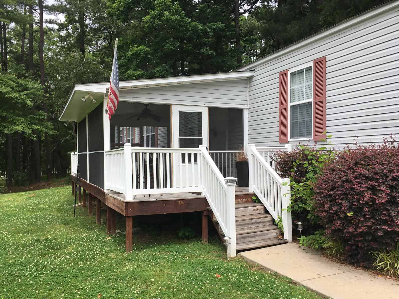 A small porch with white railings and stairs leads to the side entrance of a house. An American flag is mounted on the porch. The house has red shutters, and the surrounding yard is grassy with trees and bushes nearby.