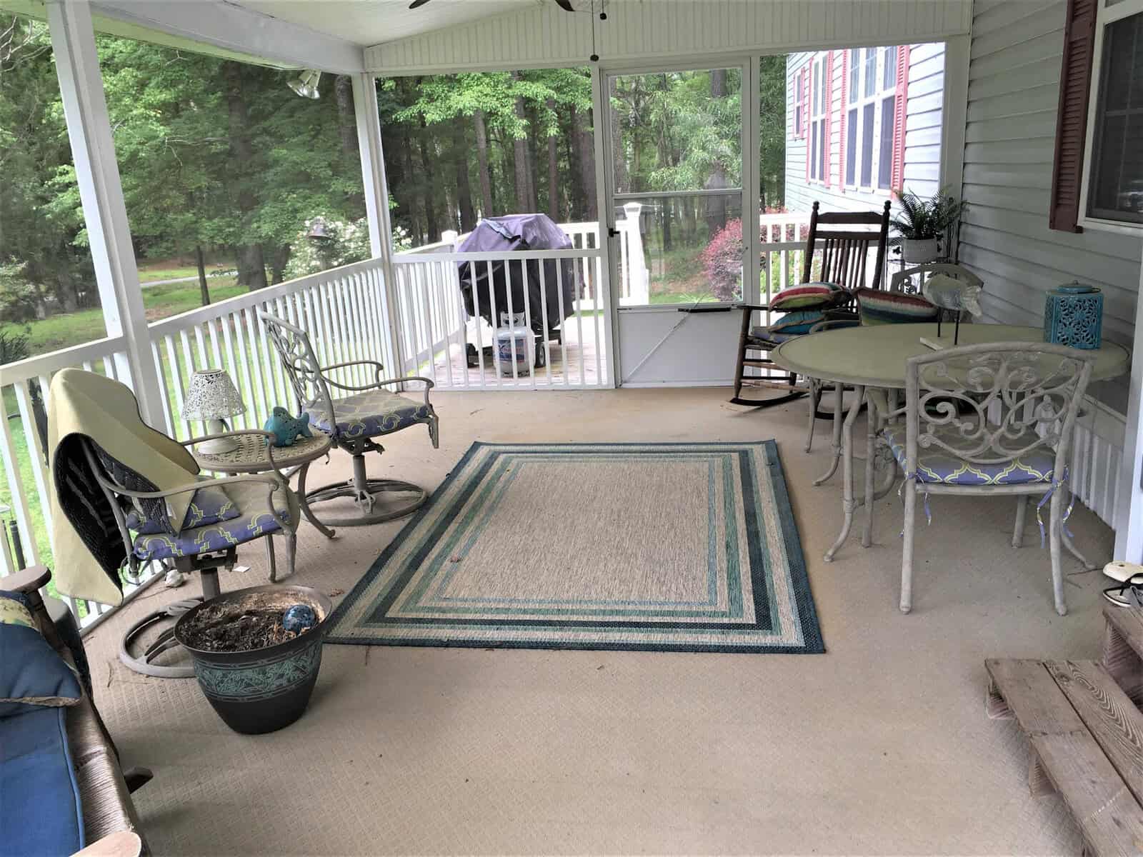 A cozy screened-in porch with white railings features a round table with chairs, a rocking chair, a few potted plants, a grill, and an outdoor rug. The porch overlooks a lush, green forested area.