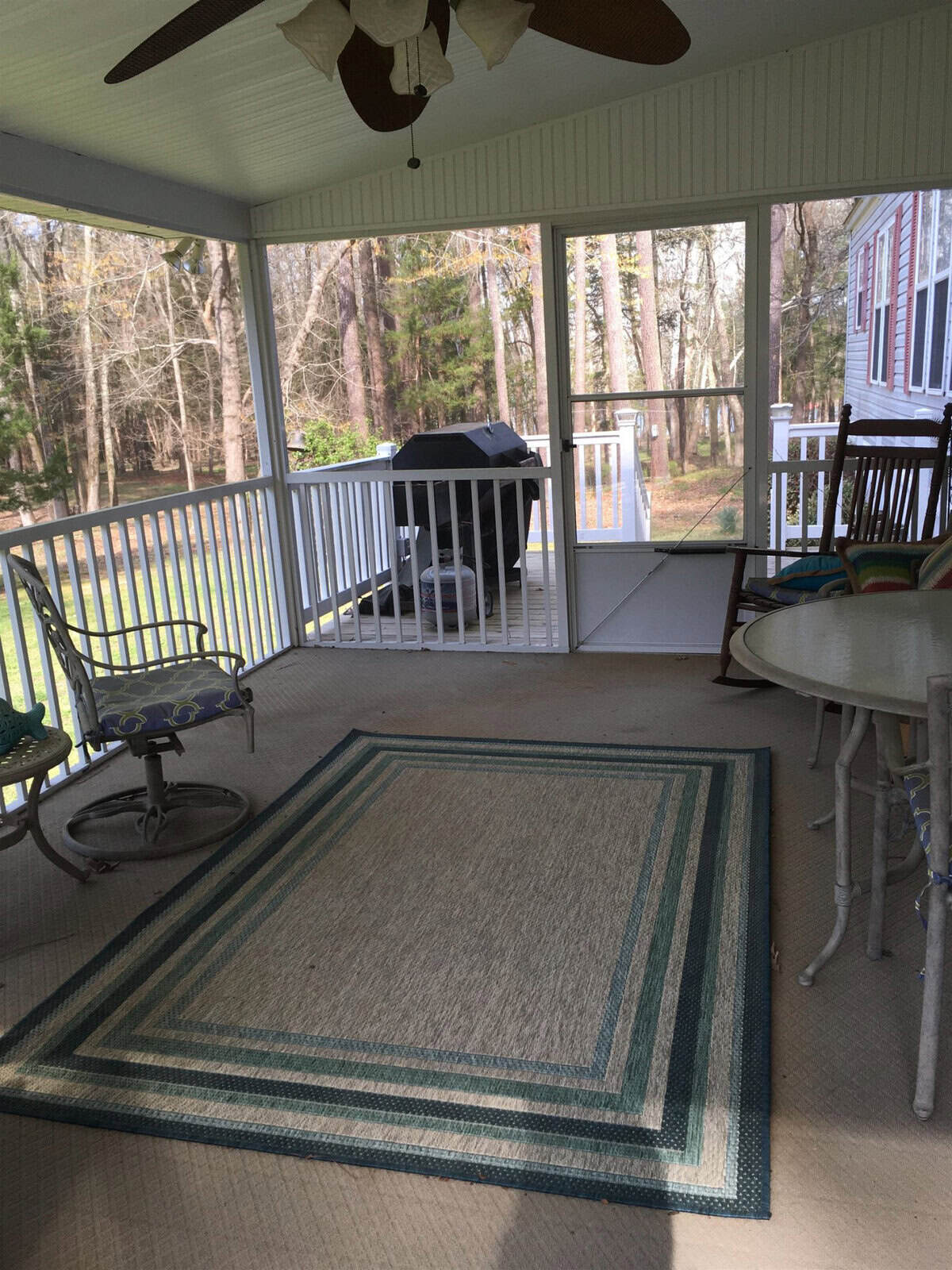 A screened porch with white railings, featuring a ceiling fan, rocking chairs, and a striped area rug. A grill is visible outside on a wooden deck surrounded by a forested area.
