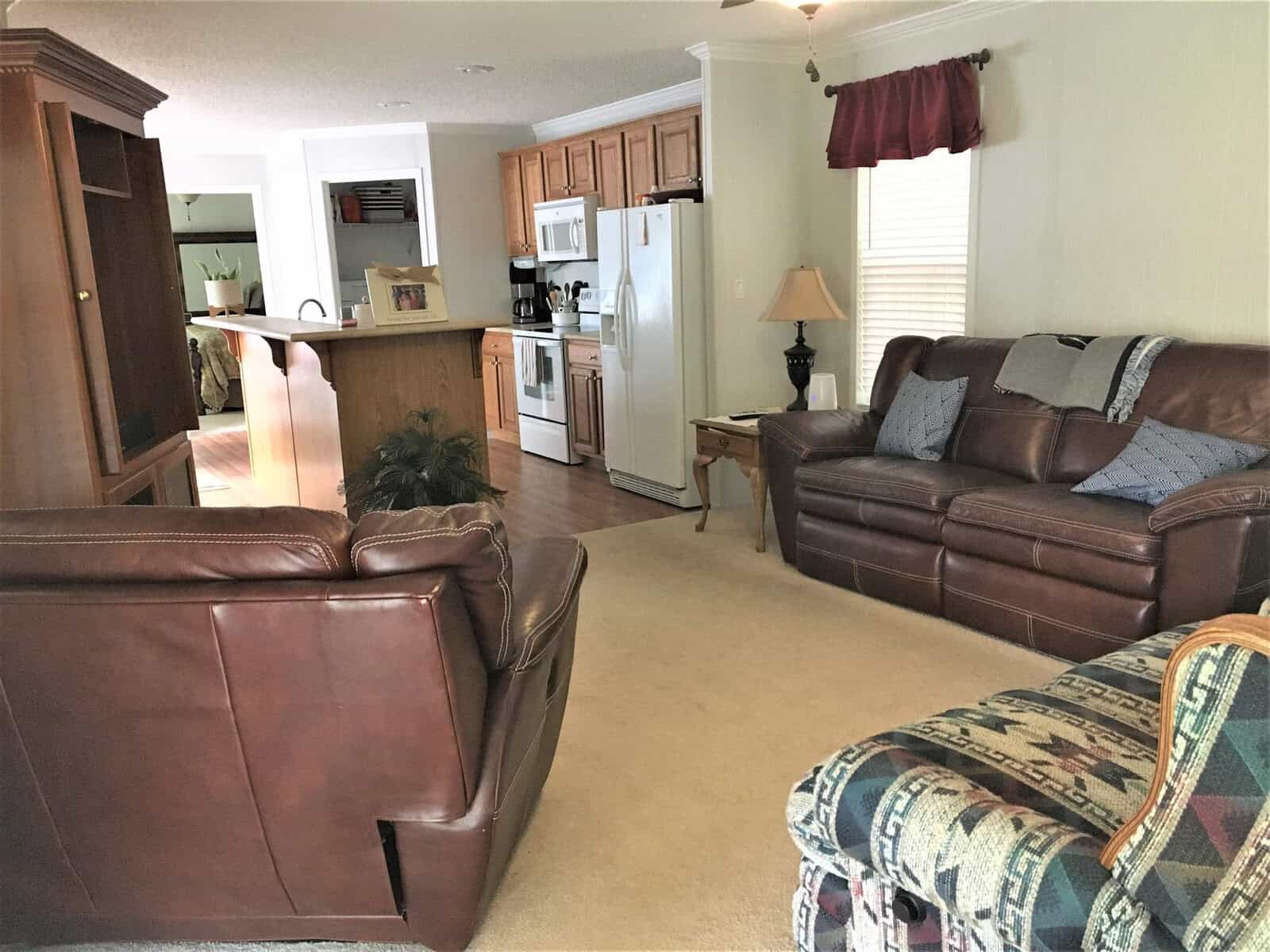 A cozy living room with a brown leather sofa set, a wooden coffee table, and a patterned armchair. The room opens to a kitchen with wooden cabinets and white appliances. A lamp is on a side table, and a carpet covers the floor.