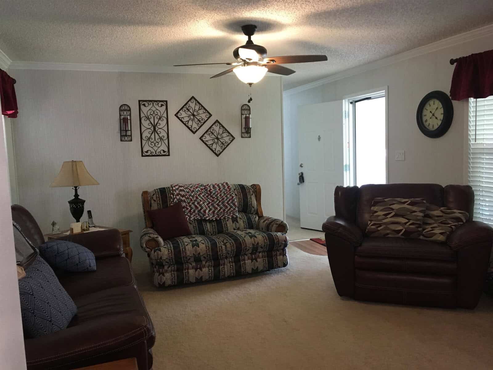 Cozy living room with a ceiling fan, two armchairs, a patterned sofa, and a floor lamp. Wall decorations include framed art, a clock, and birdcage accents. Light streams in from a window with red curtains and an open door.