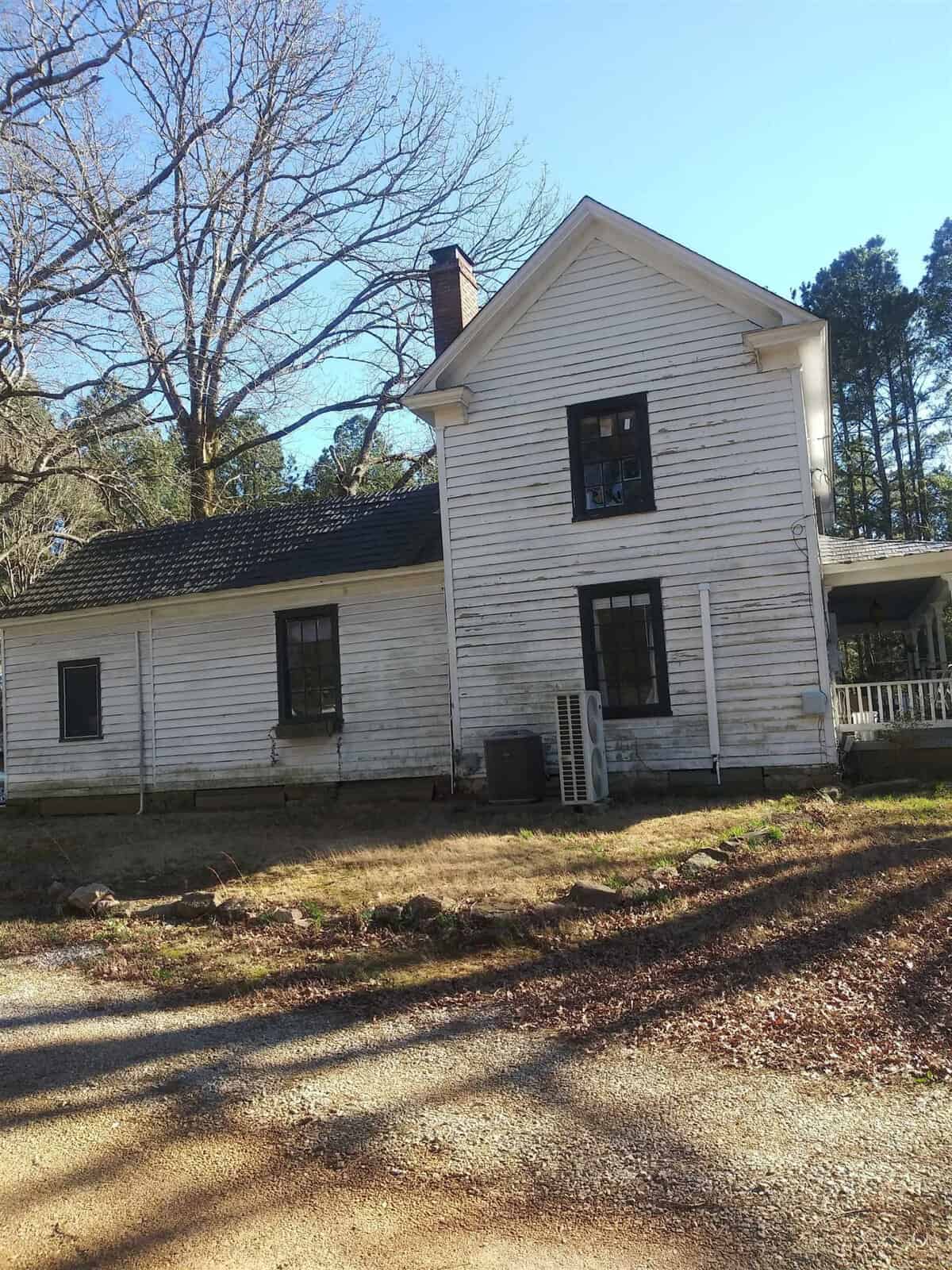 A weathered two-story white wooden house with peeling paint and a blackened window, situated next to a dirt road with surrounding bare trees, suggesting neglect or abandonment.