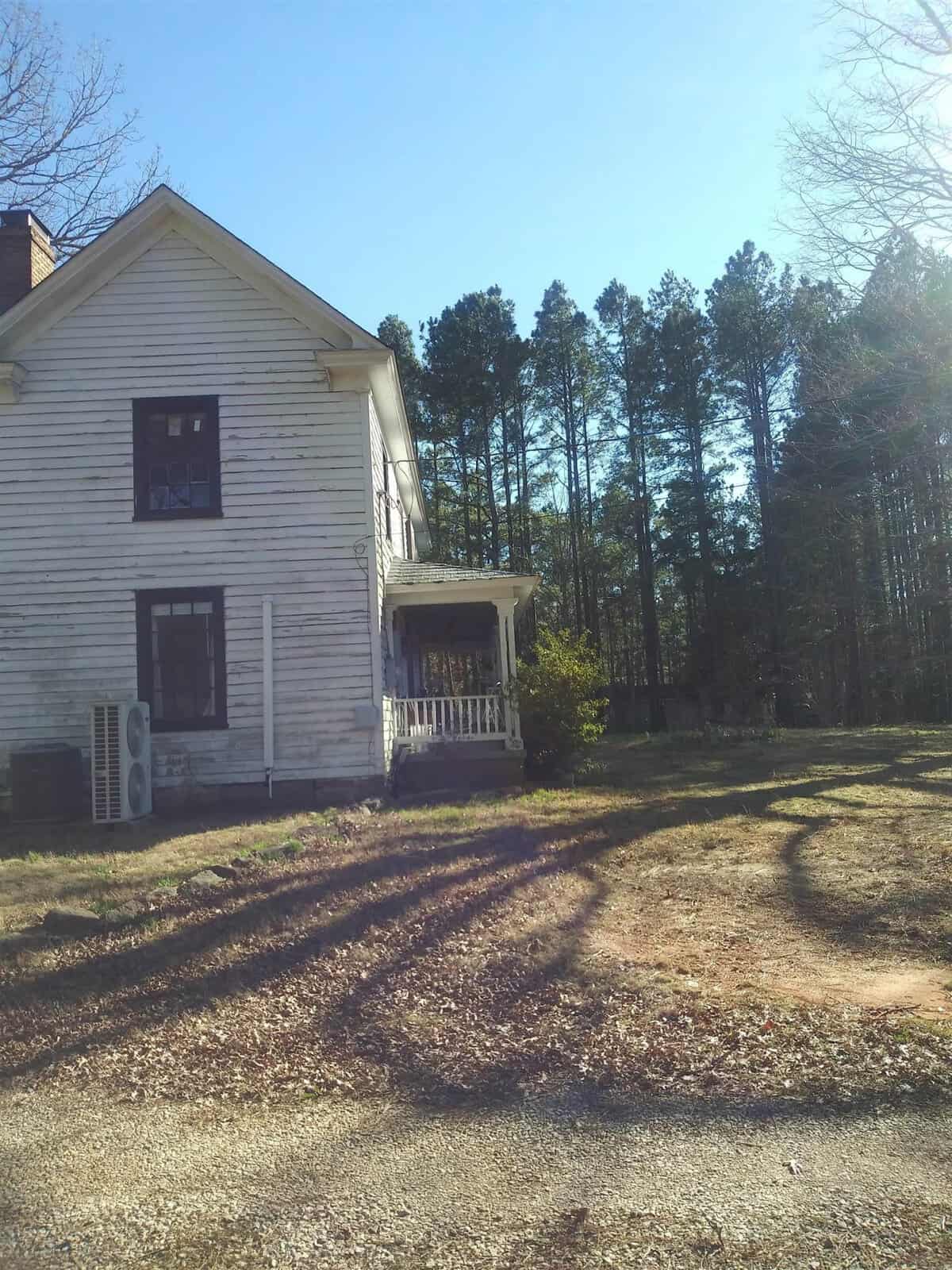 A two-story white house with a porch on a sunny day, with shadows of trees cast on the ground and surrounded by a wooded area.