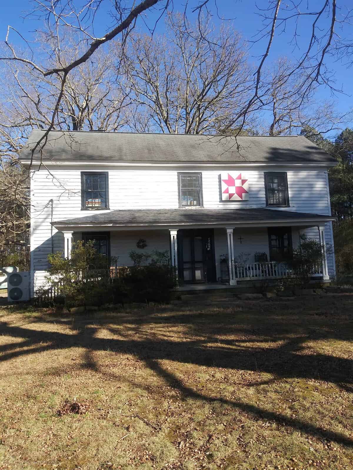 A two-story white house with dark shutters and a single pink star decoration centered above the front porch, set against a backdrop of bare trees and a clear sky.