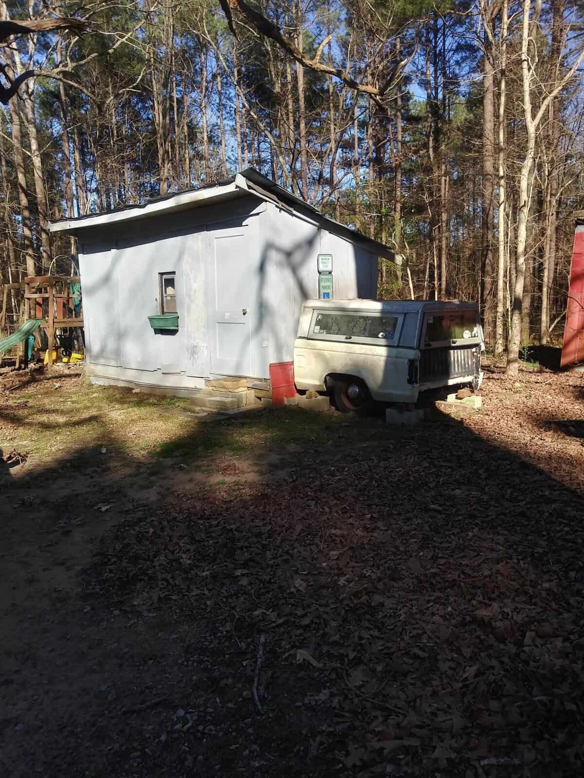 A white, vintage-style van is parked beside a small, gray building with a green door and white trim, surrounded by a wooded area with leaf-strewn ground.