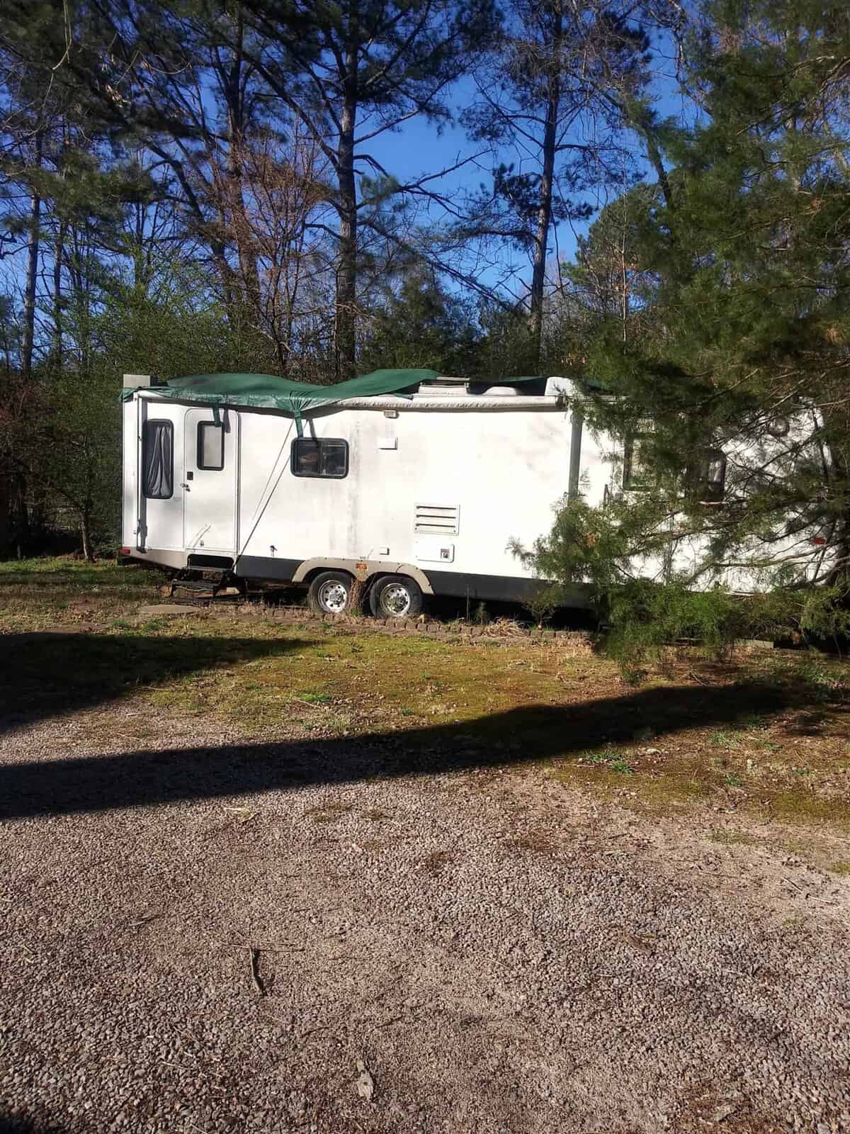 A travel trailer parked on a gravel surface surrounded by trees with an extended awning on the side.
