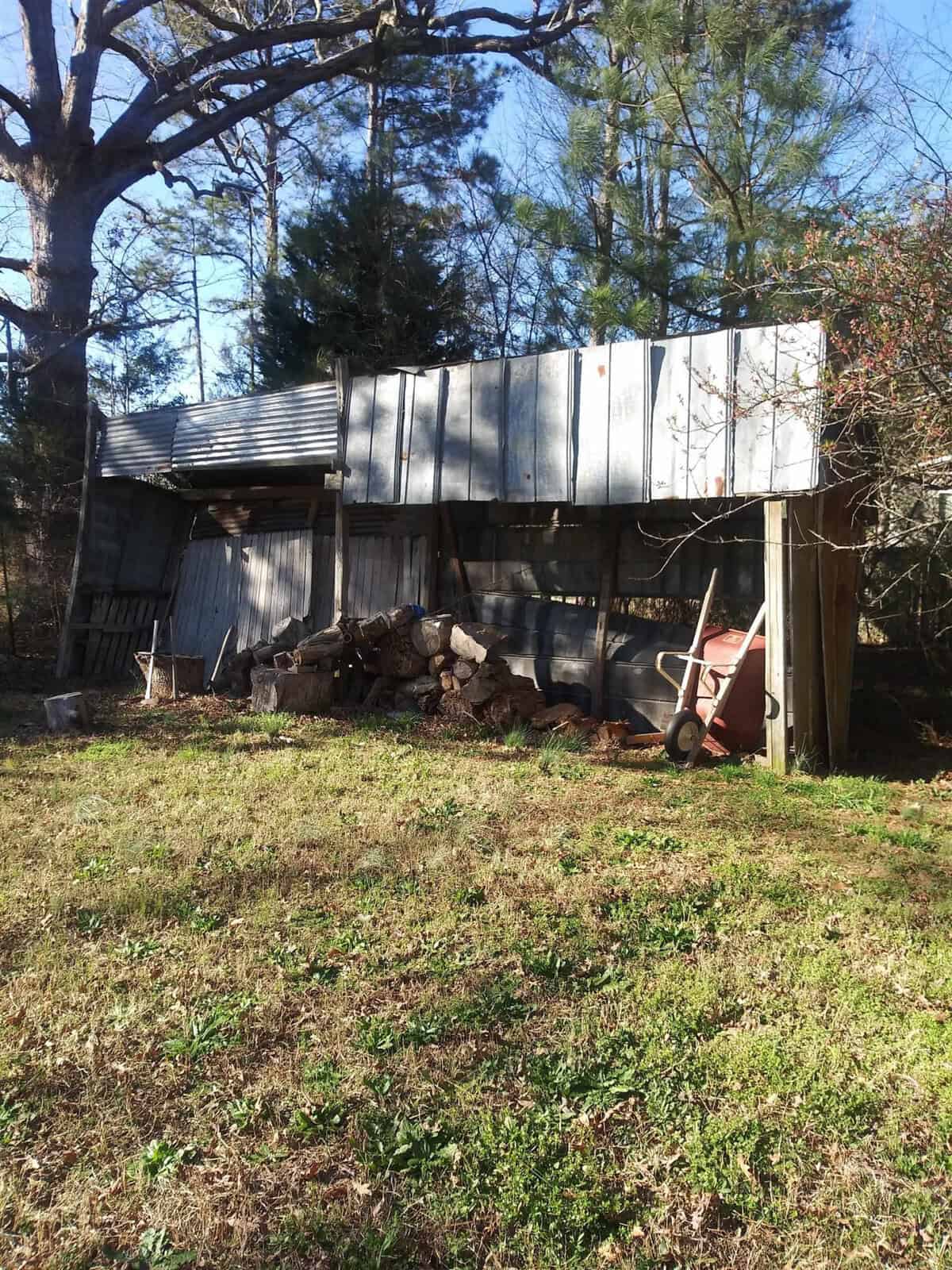 Rustic wooden shed with tin roofing storing firewood, flanked by a wheelbarrow and assorted garden tools, set against a backdrop of trees under a clear sky.